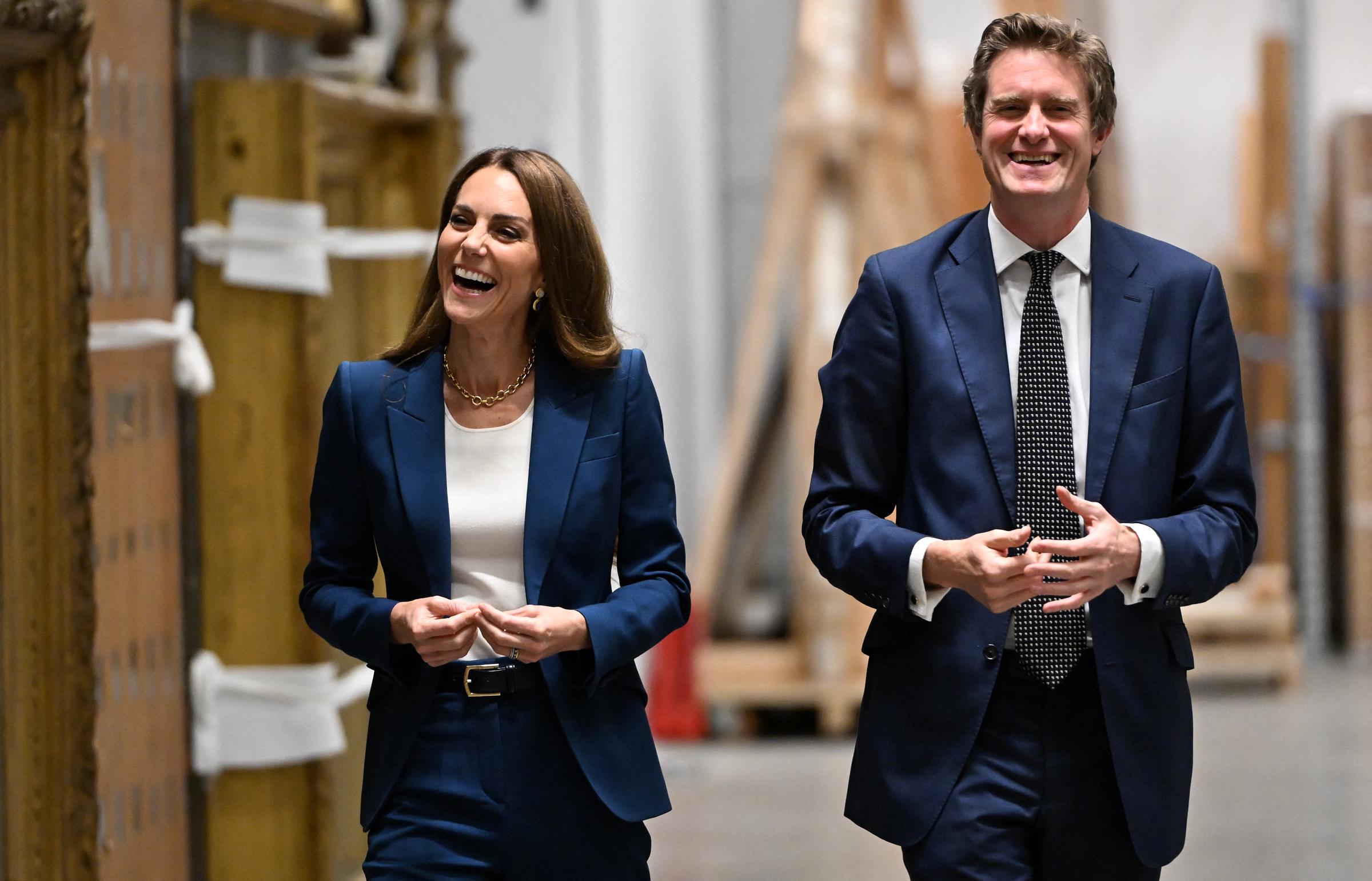 Prinzessin Catherine lacht gemeinsam mit Tristram Hunt und wirkt entspannt und engagiert. Ihr Gesichtsausdruck ist offen und aufrichtig und verleiht ihrem geschmackvollen Anzug in Marineblau und goldenen Accessoires Wärme.