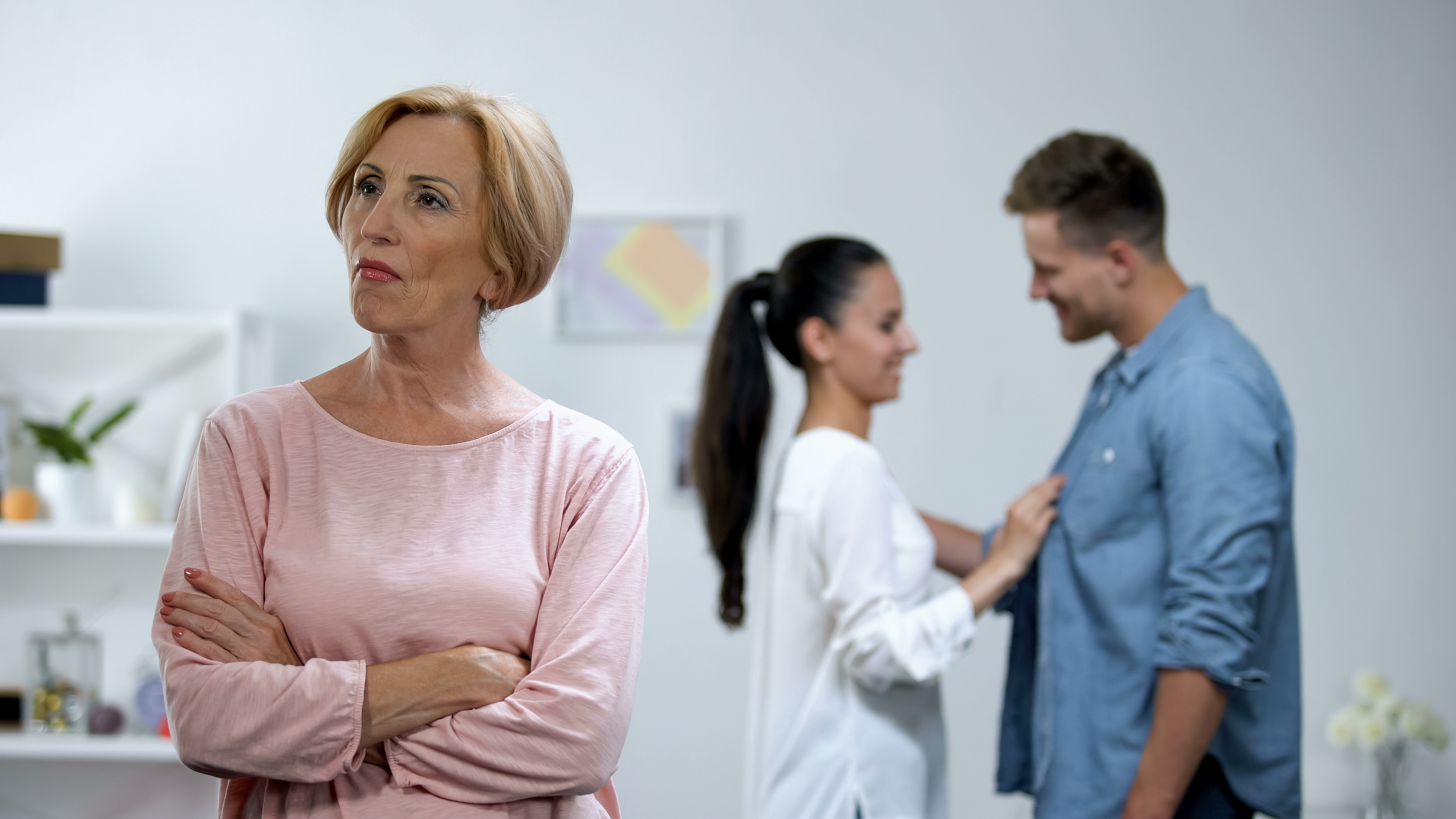 Eine Schwiegermutter steht mit verschränkten Armen vor ihrem Sohn und ihrer Schwiegertochter, die im Hintergrund glücklich wirken | Quelle: Shutterstock