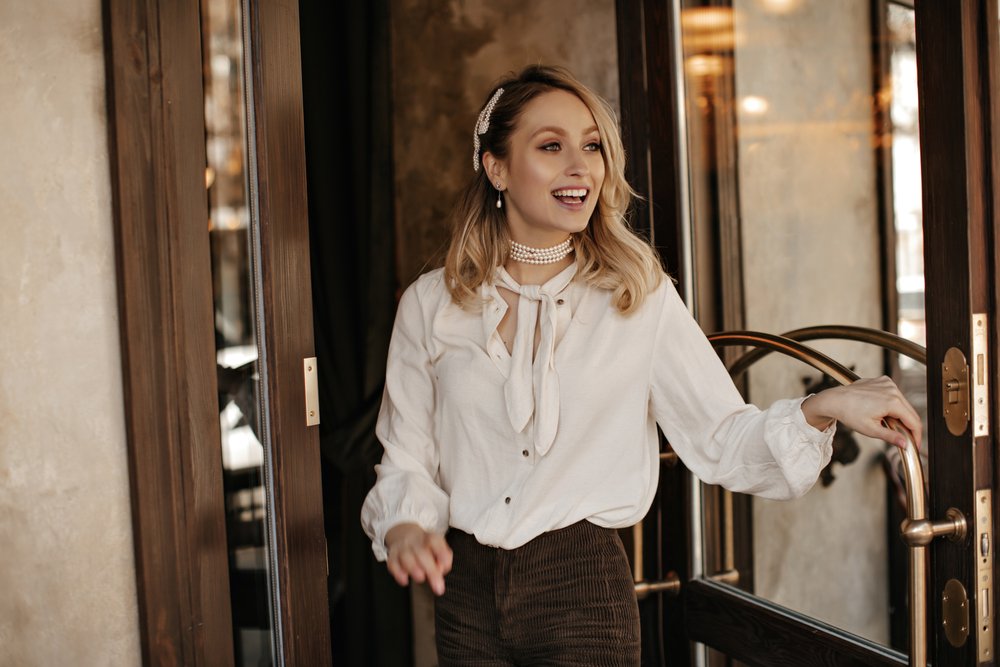 Fröhliche, lockige blonde Frau in weißer Bluse und brauner Samthose lächelt aufrichtig und öffnet Tür. I Quelle: Shutterstock