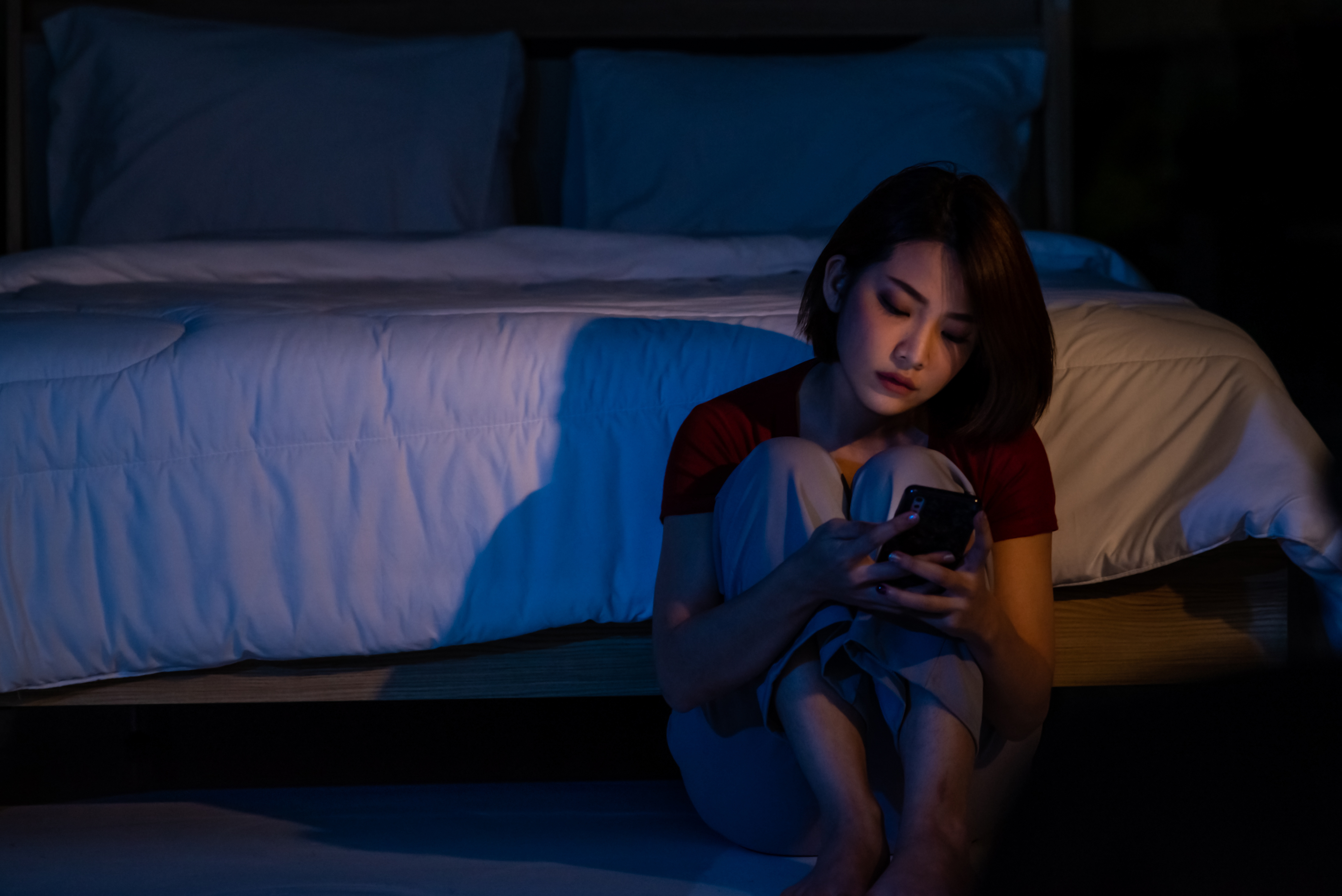 Depressive junge schöne asiatische Frau, die allein auf dem Boden im Schlafzimmer sitzt und auf ihr Smartphone schaut. | Quelle: Getty Images