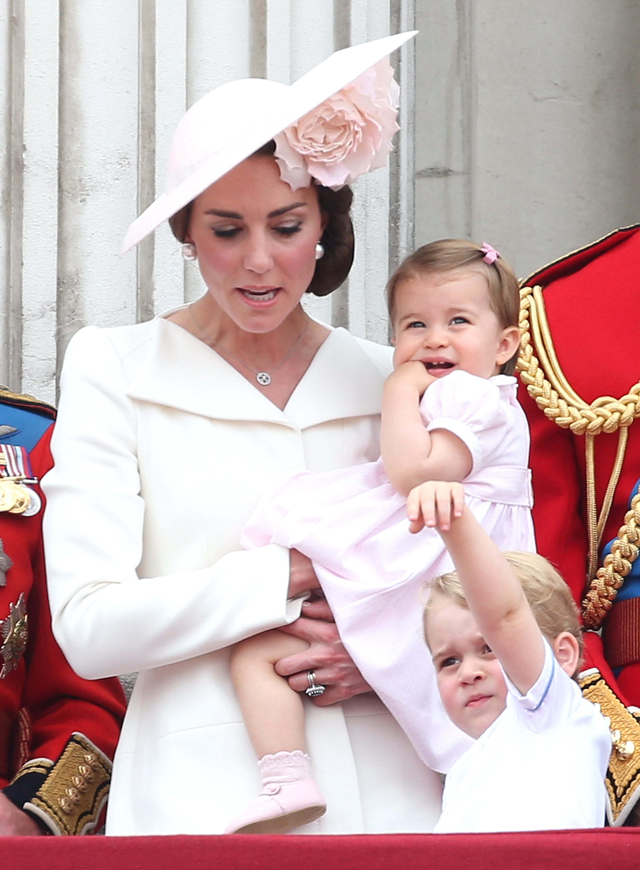 Catherine, Herzogin von Cambridge, hält die kleine Prinzessin Charlotte im Arm, während sie zusammen mit Prinz George auf dem Balkon steht – während der „Trooping the Colour“-Parade auf der Mall am 11. Juni 2016 in London, England. Catherine sieht in einem elfenbeinfarbenen, taillierten Mantelkleid und einem blassrosa Fascinator von Philip Treacy, der mit einer großen Blüte verziert ist, einfach umwerfend aus, während Charlotte ein pastellrosa Kleid mit Puffärmeln und passenden Schuhen trägt und George in einem weißen Outfit mit Kragen nach oben greift. | Quelle: Getty Images