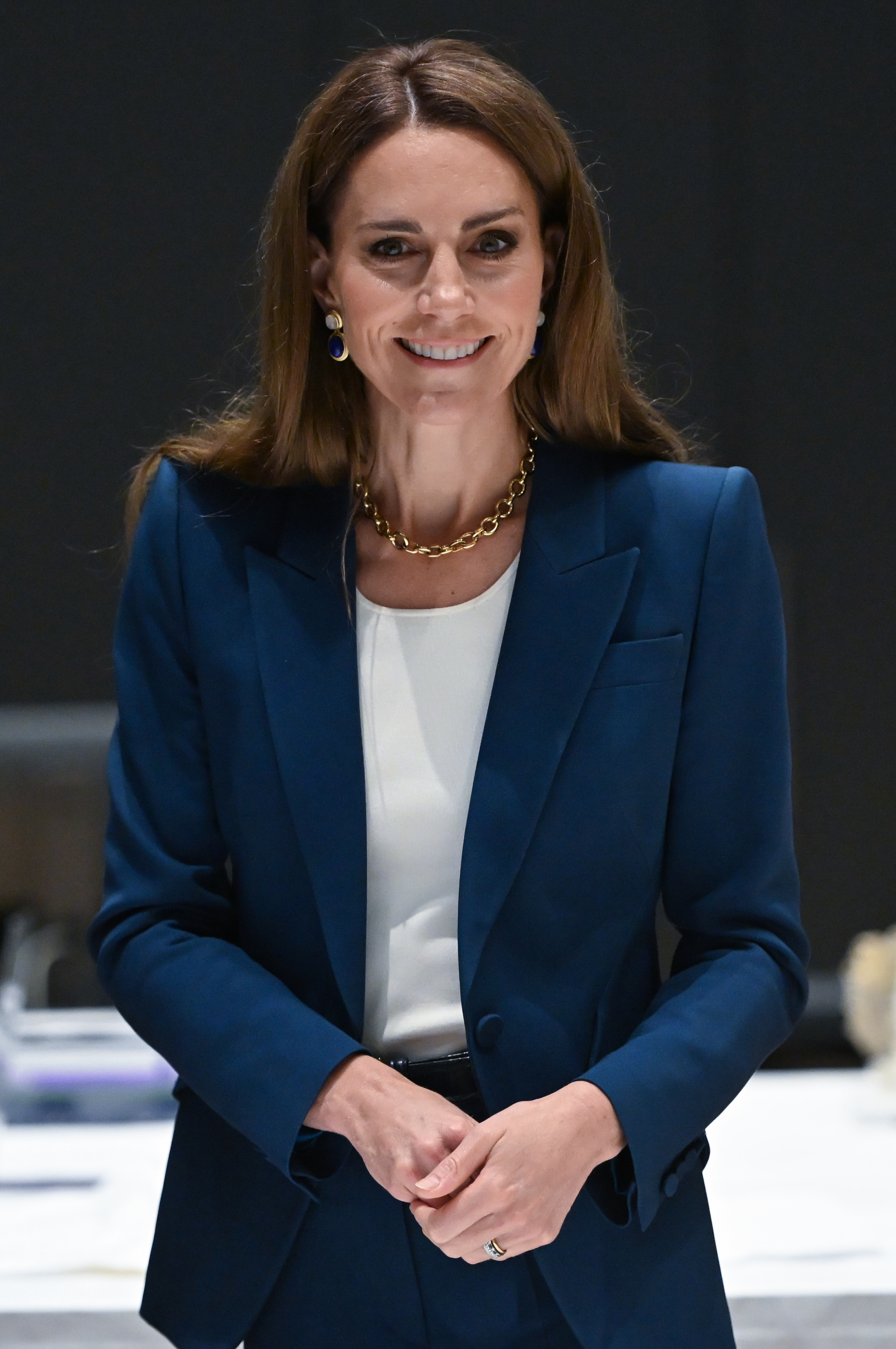 Prinzessin Catherine lächelt sanft in einem maßgeschneiderten blauen Anzug und einem weißen Oberteil. Ihre Haltung ist aufrecht, die Hände sanft gefaltet und sie strahlt ruhiges Vertrauen aus, während sie im V&A East Storehouse steht.