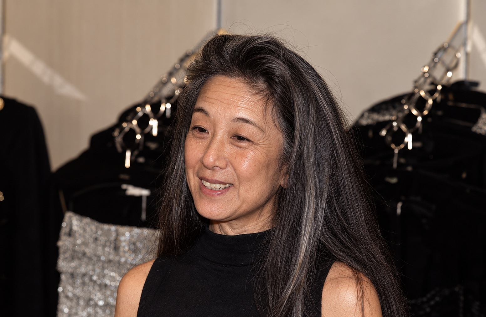 Diese KI-Kreation zeigt eine alternative Version von Vera Wang: natürliche silberne Strähnen, die sich in ihr langes dunkles Haar mischen, sichtbare Reife um die Augen herum und ein unverändertes Nasenprofil. Das simulierte Bild, das in einer Kulisse aufgenommen wurde, die an einen Backstage-Showroom erinnert, zeigt die Modeikone in einem hochgeschlossenen, ärmellosen schwarzen Oberteil mit einem entspannten, ungezwungenen Gesichtsausdruck. | Quelle: ChatGPT