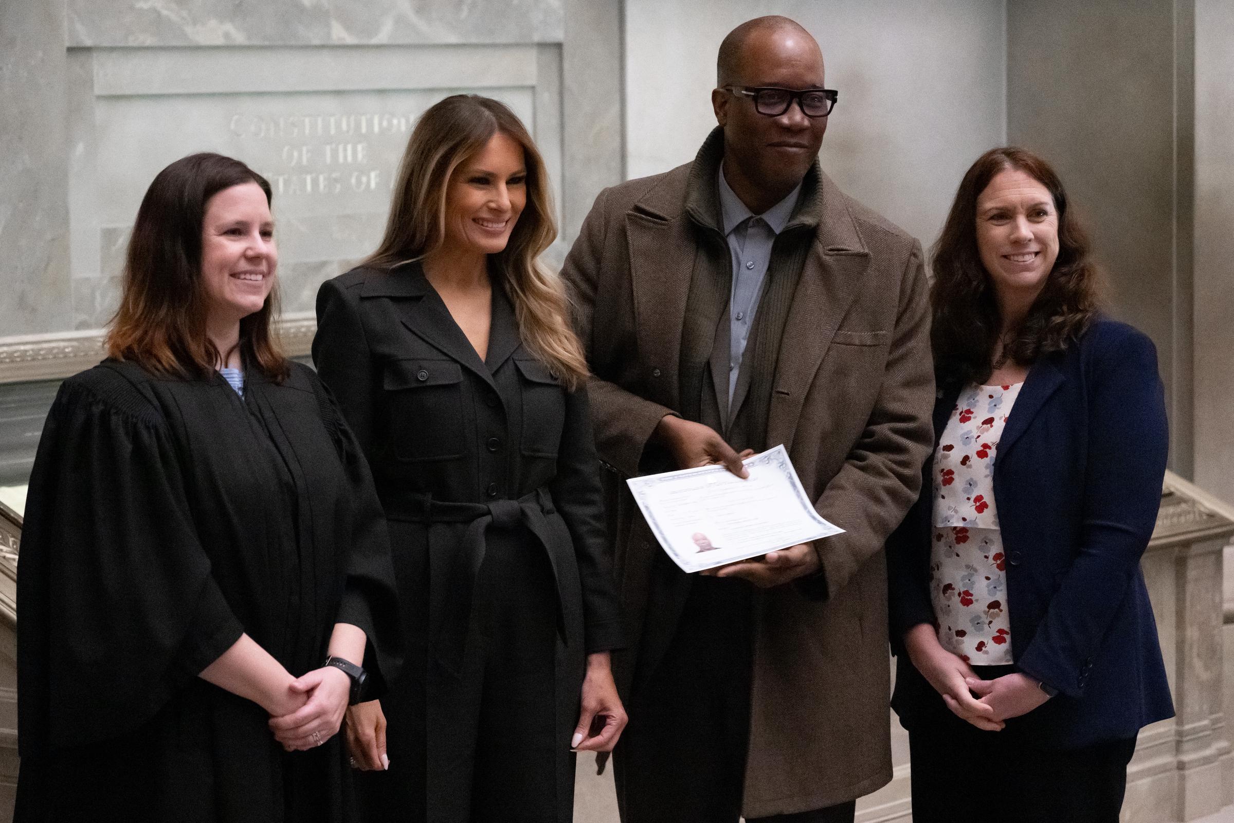 First Lady Melania Trump stand neben der US-Insolvenzrichterin Elizabeth Gunn und der Archivarin der Vereinigten Staaten, Colleen Shogan, als sie nach einer Einbürgerungszeremonie im Nationalarchiv am 15. Dezember 2023 in Washington, D.C., gemeinsam mit einem frisch vereidigten Staatsbürger posierten, der seine Urkunde in der Hand hielt. Melania Trump, die bei der Veranstaltung eine Ansprache hielt, um 25 neue Staatsbürger willkommen zu heißen, trug ein komplett schwarzes, mit einem Gürtel versehenes Mantelkleid im Hemdstil und stand neben Gunn in Richterrobe und Shogan in einem marineblauen Blazer über einem geblümten Oberteil. | Quelle: Getty Images