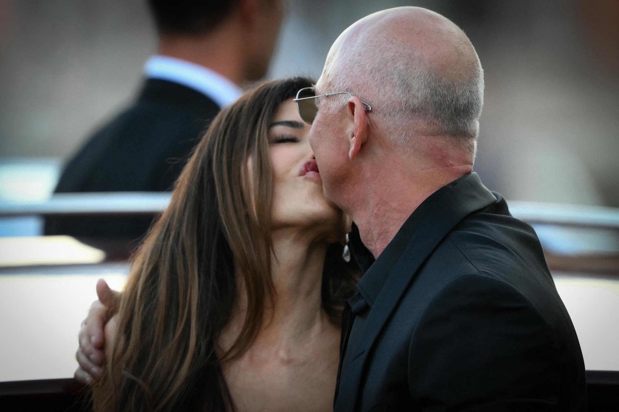 Lauren Sánchez und Jeff Bezos küssen sich, während ein Anzugträger zuschaut. Der Moment wird von verschwommenen Gebäuden und dem Abendlicht, das sich auf dem Wasser spiegelt, untermalt.
