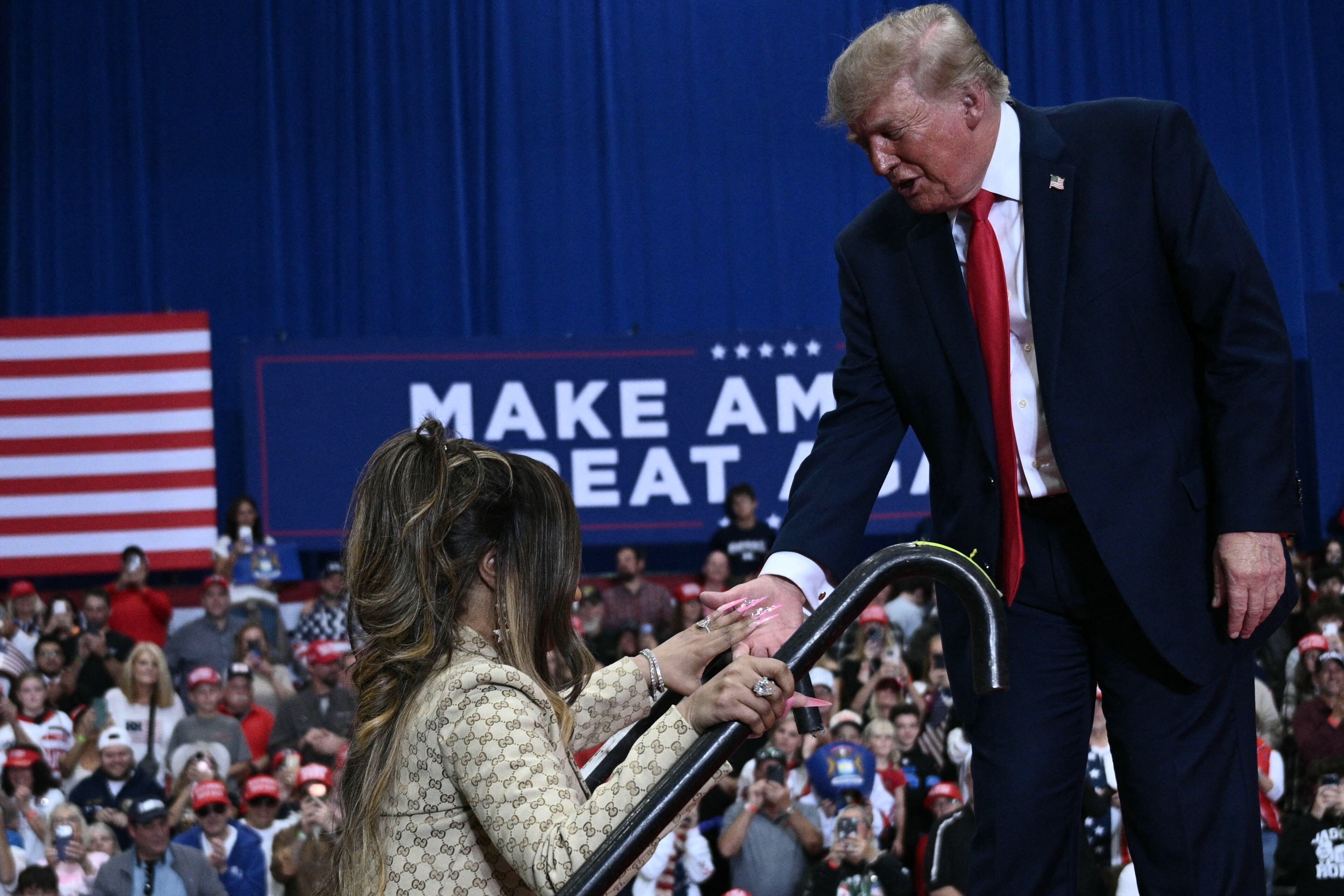 Donald Trump beugt sich vor, um Nicki Minaj auf den Stufen zur Bühne zu helfen, während sie ein Mikrofon in der Hand hält und sich am Geländer festhält – und die Zuschauer halten den Moment mit ihren Handys fest. | Quelle: Getty Images