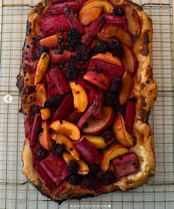 Eine frisch gebackene, rustikale Obsttorte kühlt bei Arcady auf einem Kuchengitter ab, garniert mit geröstetem Rhabarber, geschnittenen Steinobststücken und dunklen Beeren, die direkt auf dem Anwesen geerntet wurden. Diese handwerklich hergestellte Torte spiegelt Pamela Andersons kulinarische Leidenschaften und ihren pflanzenbetonten Lebensstil wider und verwandelt saisonale Gartenernten in essbare Kunst. Sie teilte diese köstliche Kreation zusammen mit dem nachdenklichen Kommentar: „Ich stelle mir das Leben wie einen Garten vor … Man kann neu pflanzen, von vorne anfangen und ihn zu seinem eigenen machen. 🤍 Liebe, P“. | Quelle: Instagram/pamelaanderson