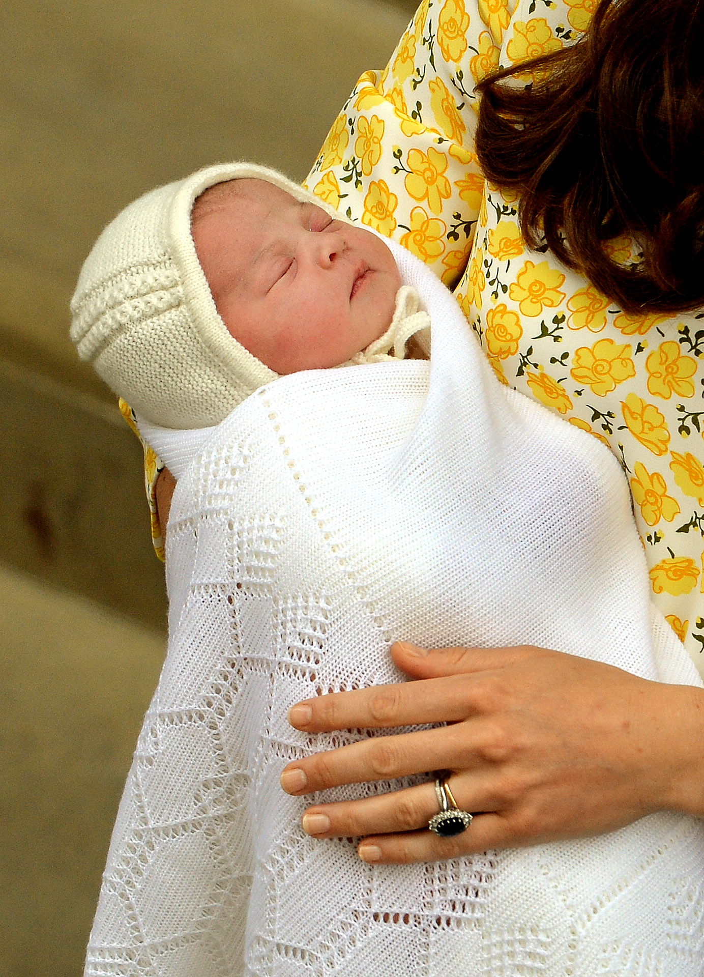 Catherine, Herzogin von Cambridge, wiegt ihre neugeborene Tochter, Prinzessin Charlotte, am 2. Mai 2015 vor dem Lindo Wing des St. Mary’s Hospital in London, England. Das schlafende Baby ist in eine weiche, weiße Strickdecke gewickelt und trägt dazu ein passendes Mützchen. Catherine hält es sicher im Arm; sie trägt ein gelbes Kleid mit Blumenmuster und ihren Saphir-Verlobungsring. | Quelle: Getty Images