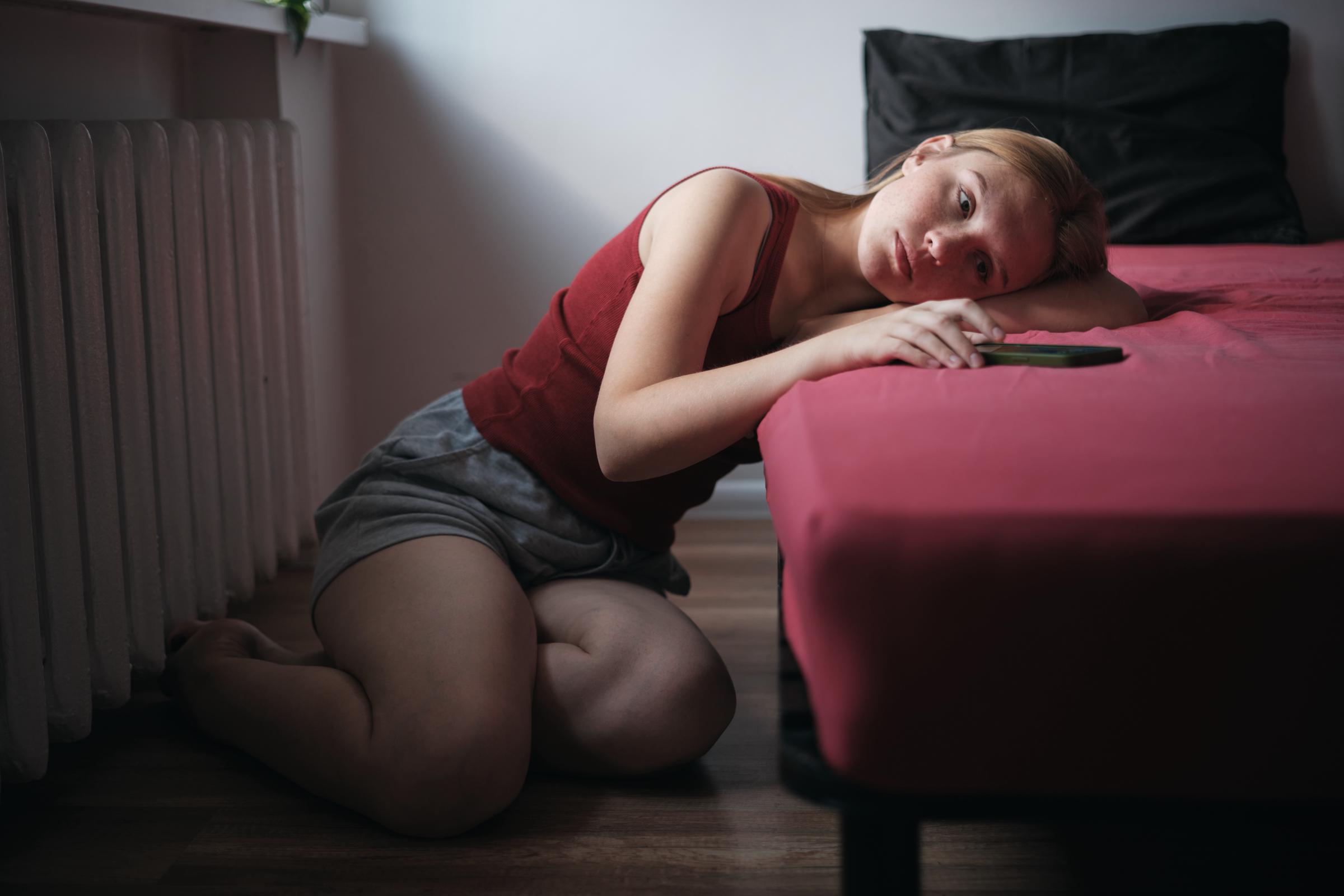 Eine deprimiert aussehende Frau, die auf dem Boden neben ihrem Bett sitzt | Quelle: Getty Images