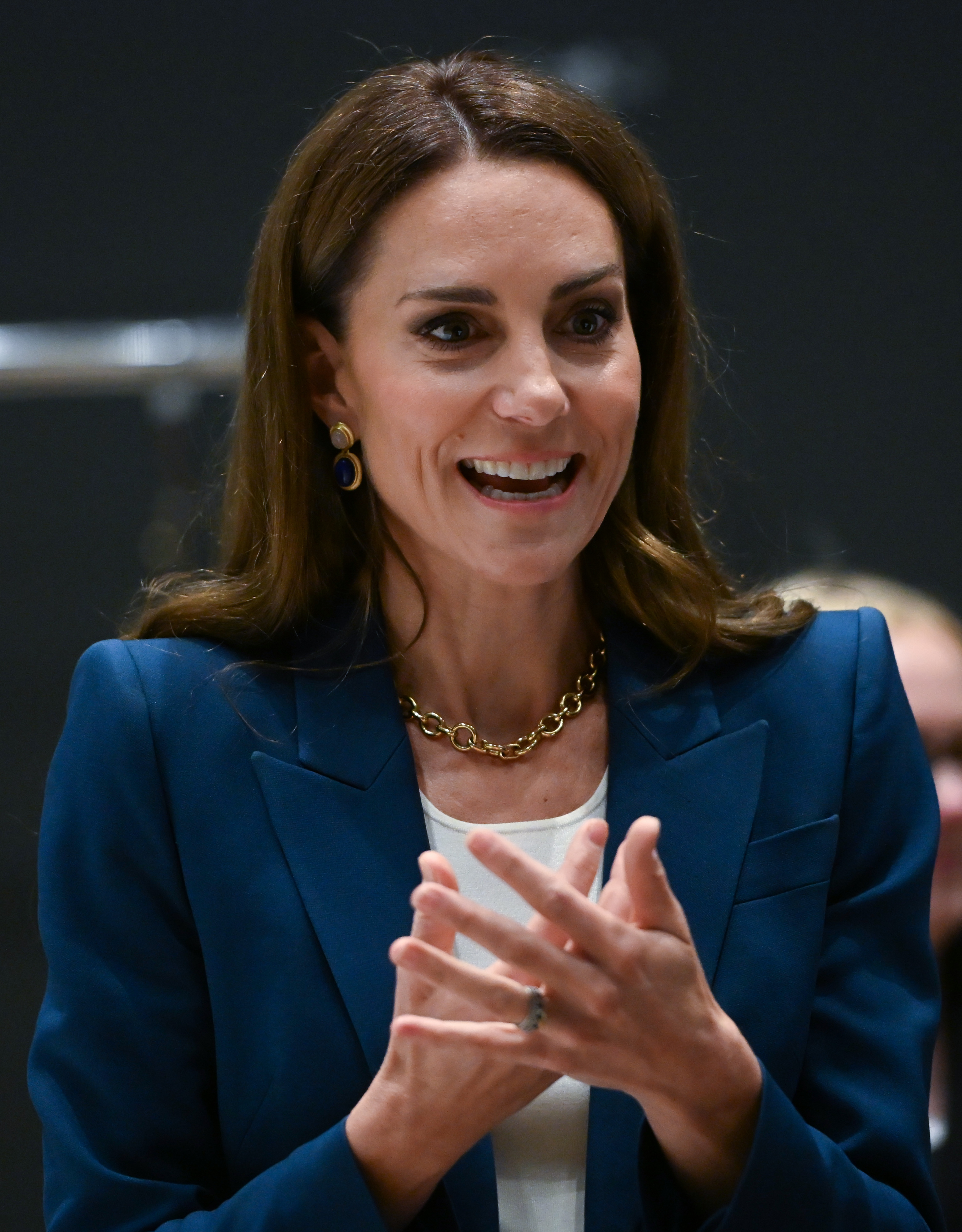 Prinzessin Catherine gestikuliert während des Gesprächs und wirkt dabei lebhaft und engagiert. Sie trägt einen kobaltblauen Blazer, eine goldene Halskette und blaue Ohrringe. Ihre elegante Frisur und ihr warmer Gesichtsausdruck unterstreichen ihre Präsenz.