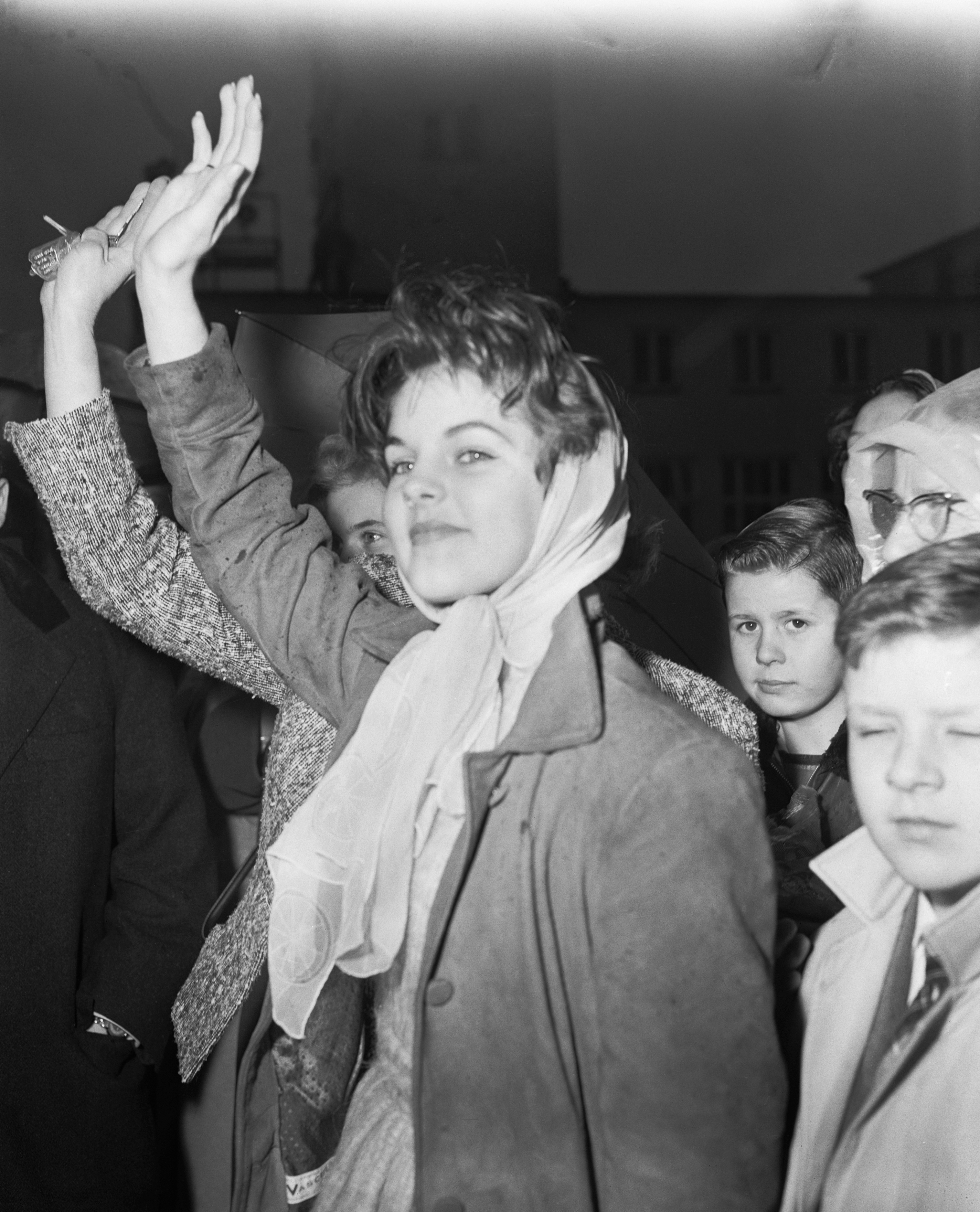 Die vierzehnjährige Priscilla Beaulieu hebt die Hand, um ihrem Freund Elvis Presley zum Abschied zu winken, als er am 2. Februar 1960 von der Rhein-Main-Air-Base in Frankfurt, Deutschland, am 2. Februar 1960, um nach 18 Monaten Dienst in der US-Armee in die Vereinigten Staaten zurückzukehren. Sie trägt einen maßgeschneiderten Mantel und ein helles Kopftuch, das locker unter ihrem Kinn gebunden ist. | Quelle: Getty Images