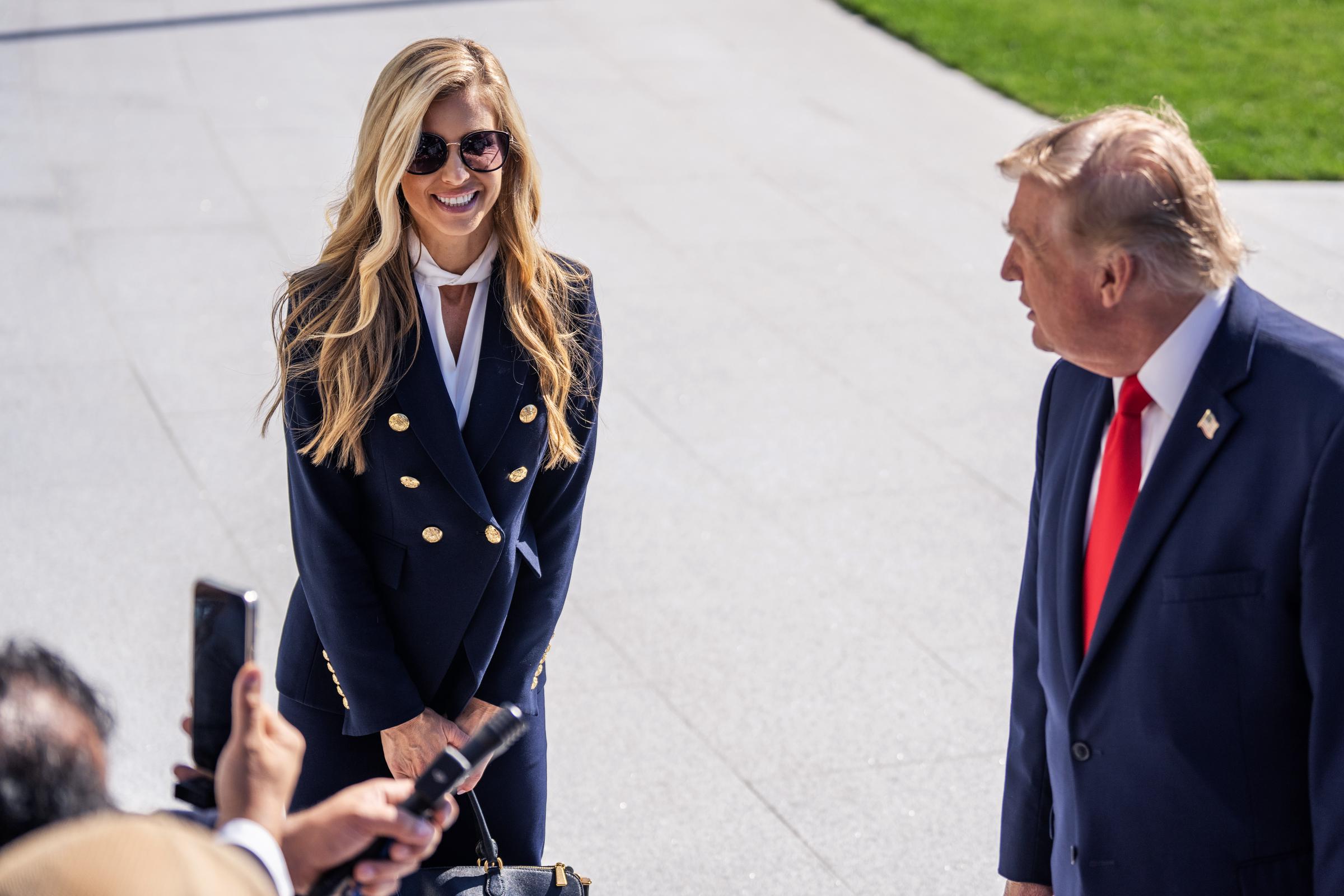 Die Pressesprecherin des Weißen Hauses, Katie Zacharia, und Präsident Donald Trump sprechen am 13. Oktober 2026 in Washington, D.C., auf dem South Lawn des Weißen Hauses mit Reportern, bevor sie an Bord der „Marine One“ abfliegen. Zacharia lächelt in ihrem marineblauen Zweireiher mit goldenen Knöpfen, einer dunklen Sonnenbrille und einer schwarzen Handtasche und steht aufmerksam neben Trump, während die Pressevertreter ihre Mikrofone und Handys zücken, um ihre Äußerungen festzuhalten. | Quelle: Getty Images