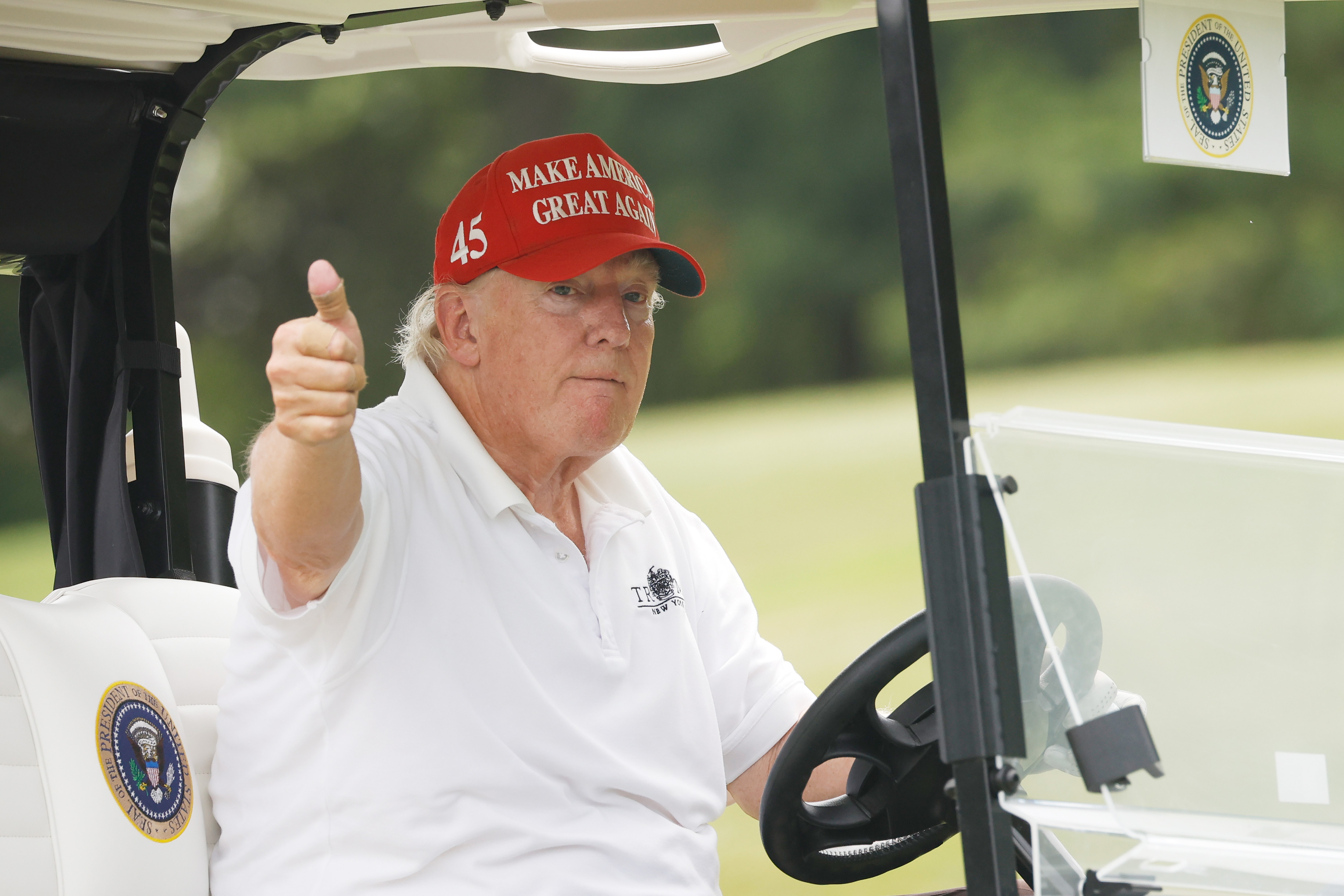 Donald Trump sitzt in einem Golfwagen und zeigt mit ernstem Gesichtsausdruck den Daumen nach oben. Der Farbunterschied zwischen seinem Gesicht und seinem Hals ist im Tageslicht sichtbar.