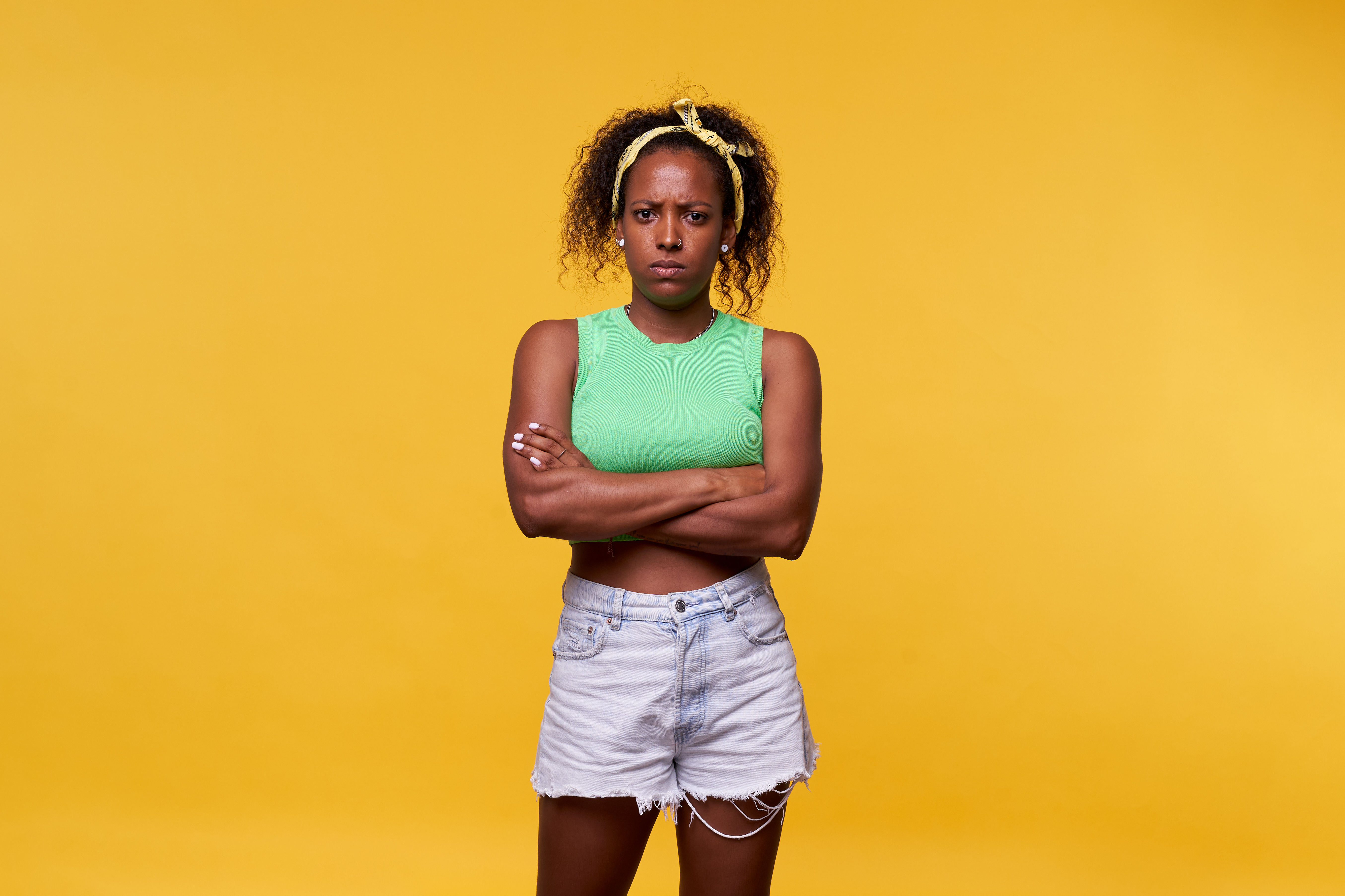 Foto einer wütenden lateinamerikanischen Frau, die vor einem gelben Hintergrund steht, die Arme verschränkt und ernst in die Kamera schaut. Einzelbild im Studio | Quelle: Getty Images