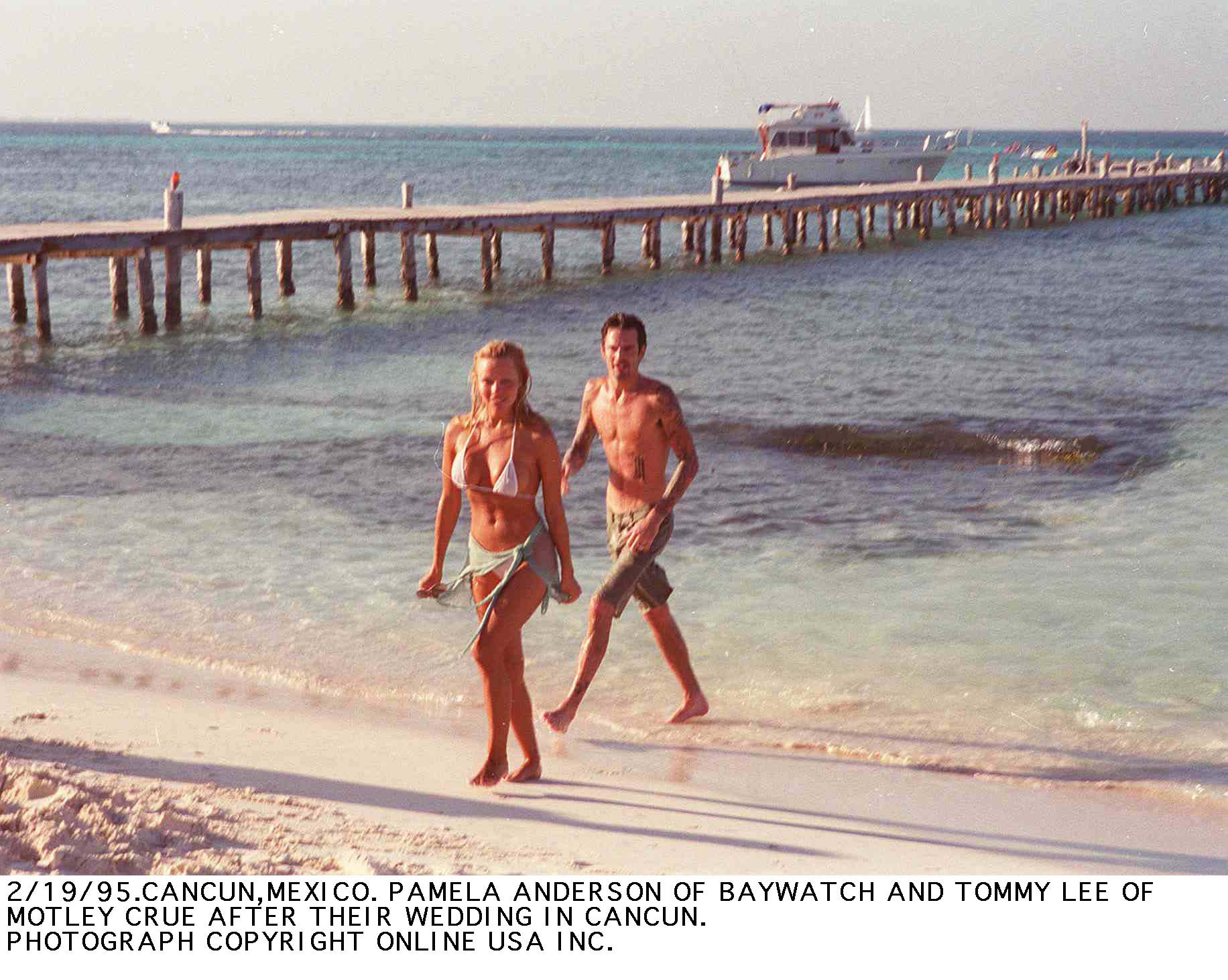 Die frischvermählten Pamela Anderson und Mötley-Crüe-Schlagzeuger Tommy Lee schlendern barfuß am sonnigen Strand von Cancún in Mexiko entlang, nur wenige Tage nach ihrer Blitzhochzeit im Februar 1995. Sie trägt einen weißen Bikini mit einem umgebundenen Sarong, während Lee Boardshorts trägt – dieser vielbeachtete Strandmoment steht symbolisch für den heftigen Medienrummel um Andersons frühen Ruhm – ein chaotisches Kapitel, das sie schließlich dazu inspirieren sollte, sich in die ruhige, bodenständige Einsamkeit von Vancouver Island zurückzuziehen. | Quelle: Getty Images