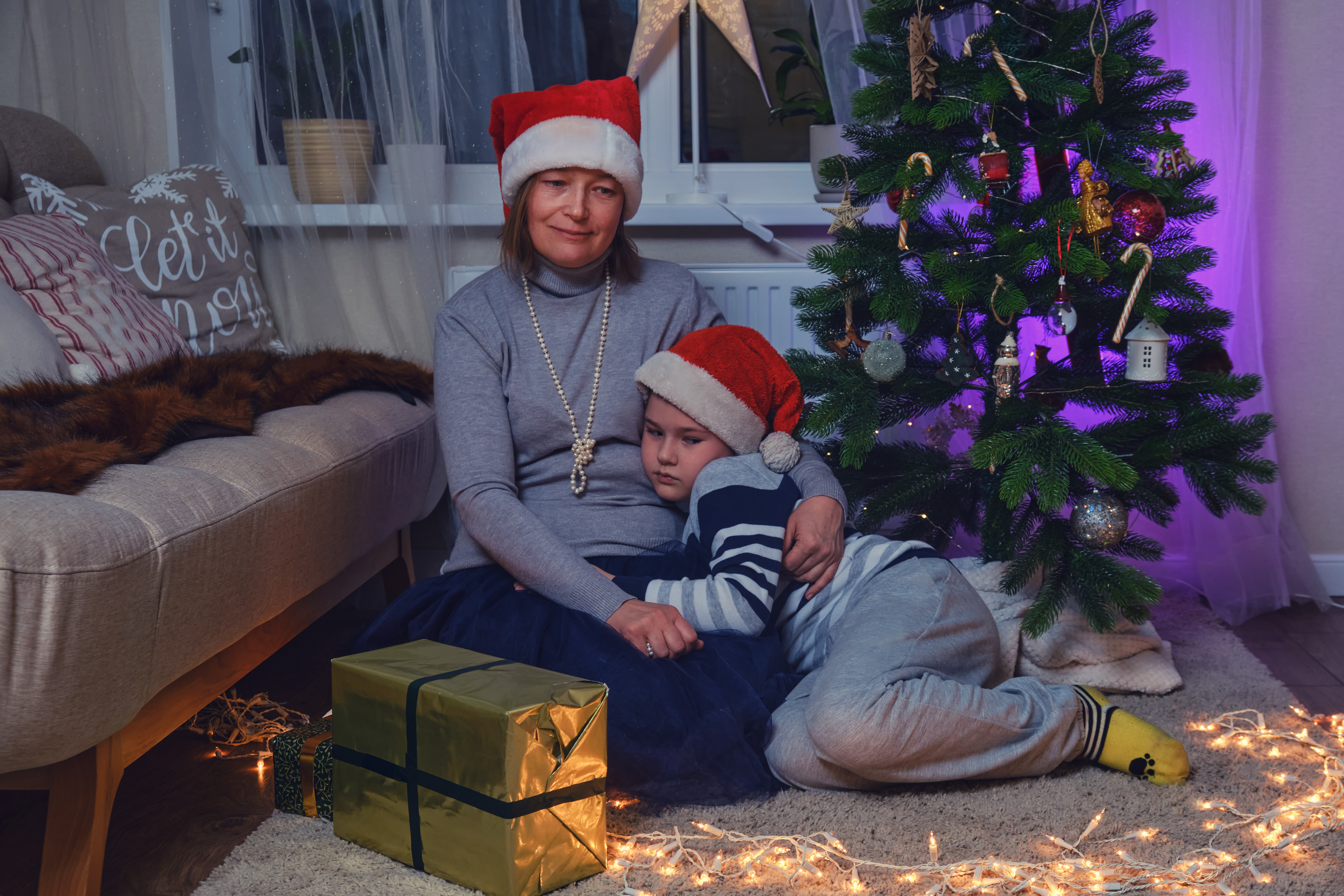 Traurige Mutter und Sohn sitzen in der Nähe des Weihnachtsbaums in den nächtlichen Lichtergirlanden. | Quelle: Getty Images