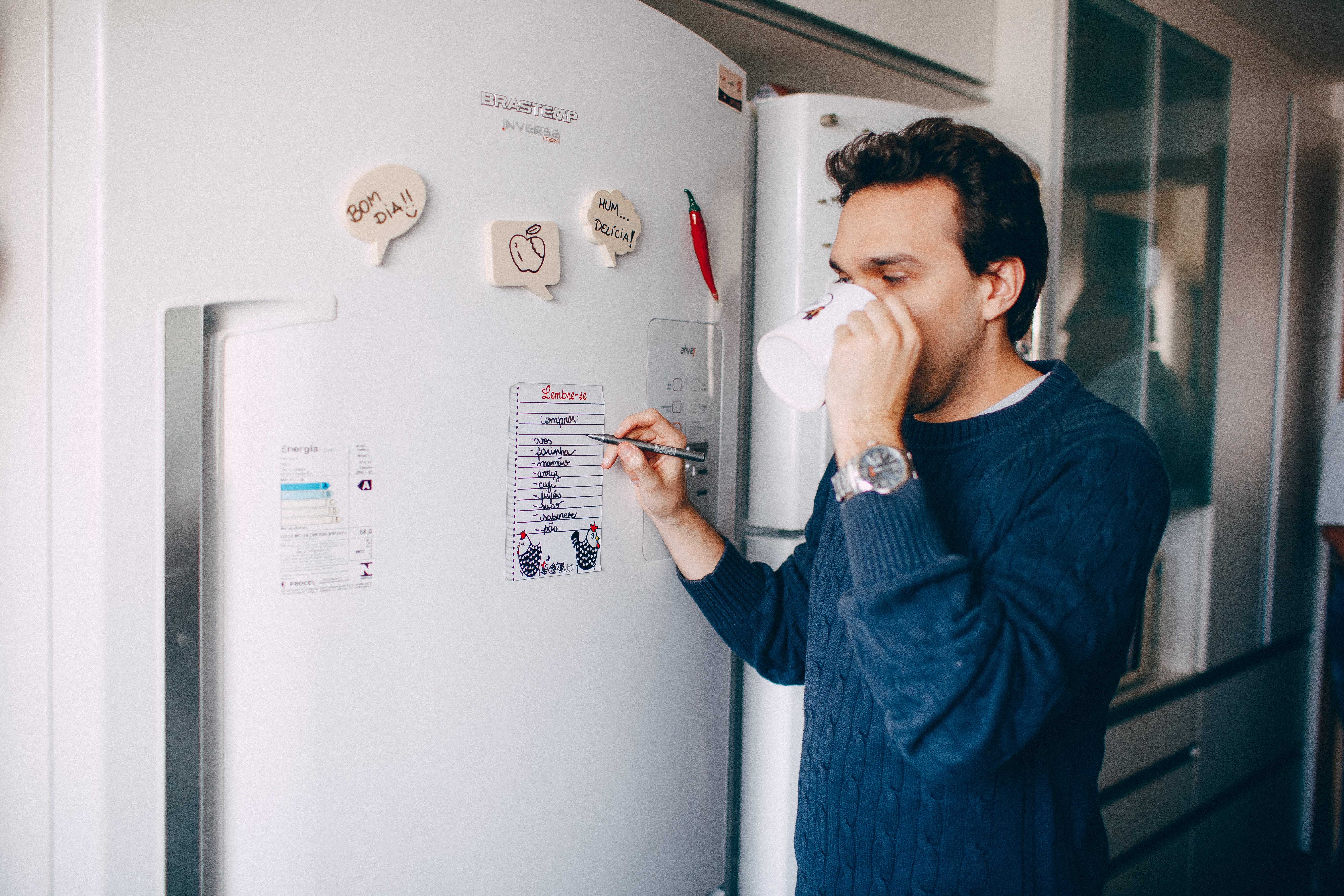 Ein Mann trinkt aus seinem Becher, während er etwas auf einen Zettel schreibt, der an einem Kühlschrank klebt | Quelle: Pexels