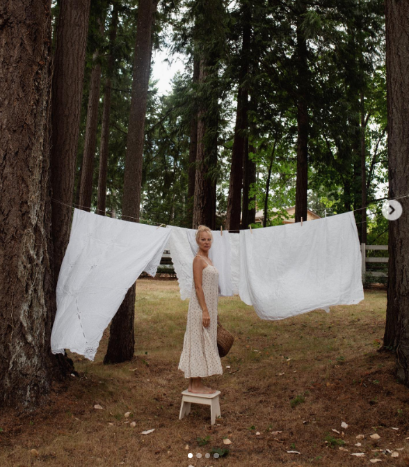 Barfuß auf einem weißen Holzhocker stehend hält Pamela Anderson kurz inne, während sie in Arcady Wäsche zwischen hoch aufragenden Nadelbäumen aufhängt. In einem zarten Sommerkleid mit Blumenmuster und hochgestecktem Haar blickt sie in die Kamera, umrahmt von flatternden weißen Wäschetüchern, und kommentiert dieses idyllische Ritual: „Ich freue mich auf die morgendlichen Aufgaben … Ich füttere und gehe mit den Hunden spazieren, ich mache die Wäsche, ich schreibe … Ich spüre, wie sich mein Leben bescheiden um mich herum entfaltet. Alles ist gut 🤍“. | Quelle: Instagram/pamelaanderson