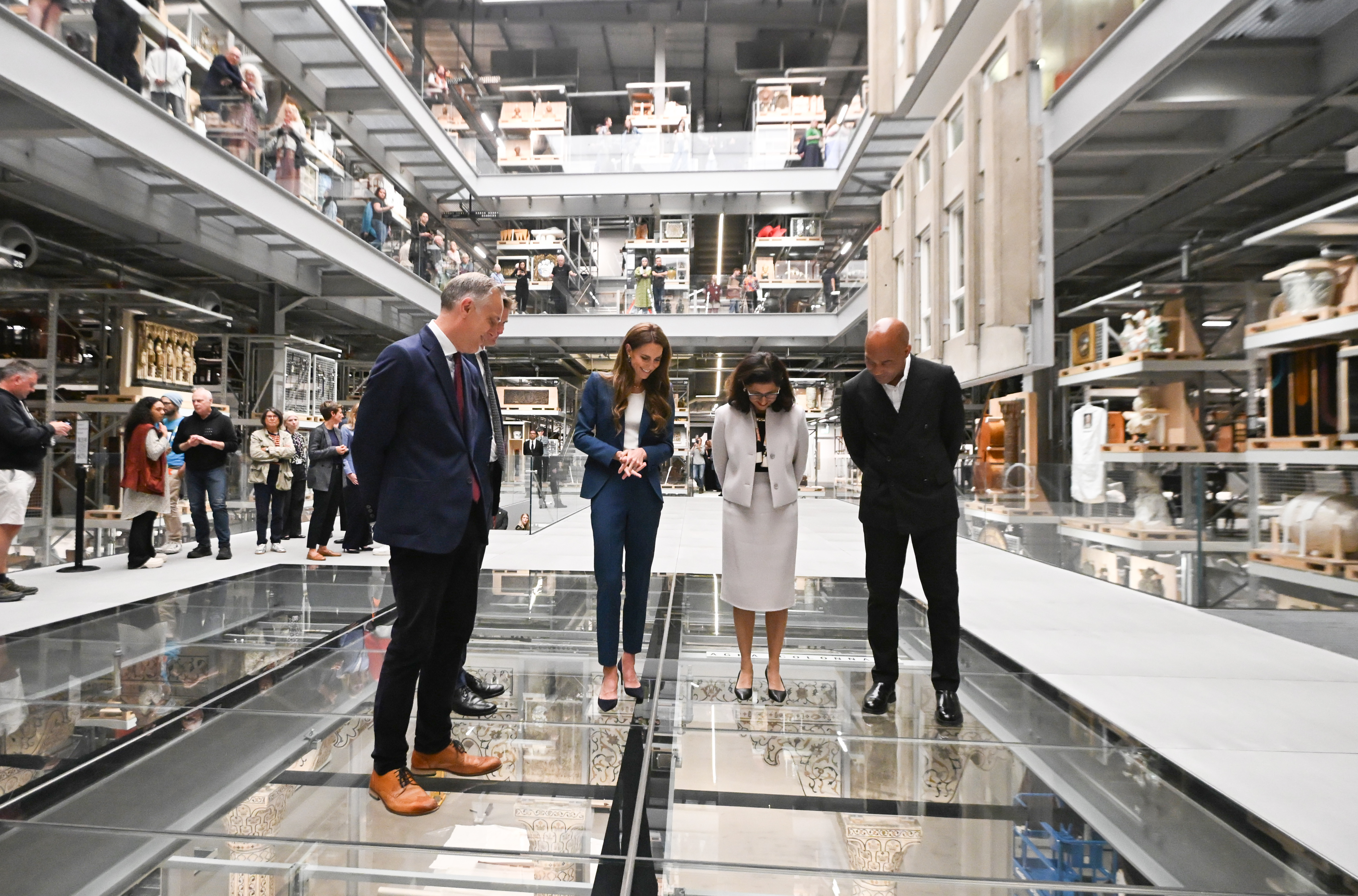 Über einem Glasboden stehend, betrachtet Prinzessin Catherine zusammen mit anderen Museumsführern die Ausstellung darunter. Ihr Blick ist nach unten gerichtet, ihre Haltung vorsichtig, was auf Neugier und Vorsicht schließen lässt, während sie den vertikalen Ausstellungsraum erkunden.