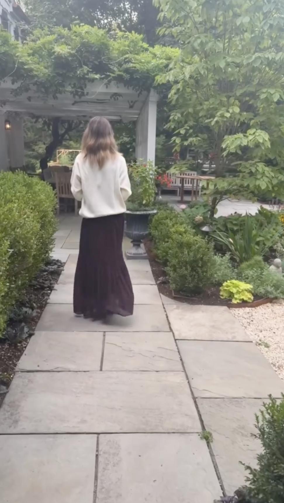 Alejandra Silva spaziert auf einem steinernen Gartenweg entlang, der von gepflegten Hecken und blühenden Pflanzen gesäumt ist, am Haus ihrer Familie in Spanien. | Quelle: Instagram/alejandragere