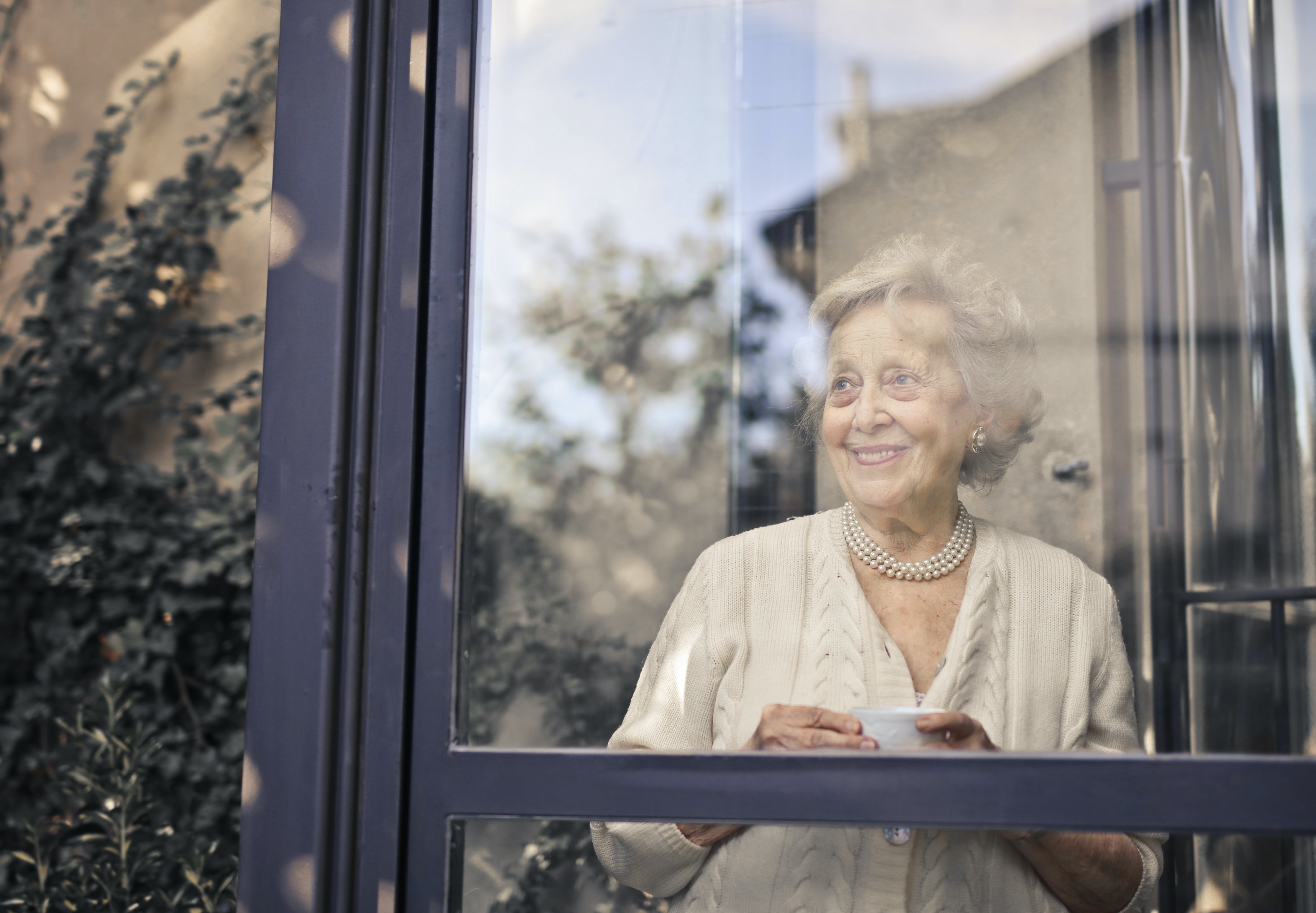 Eine ältere Frau lächelt, während sie ein Getränk in der Hand hält und aus dem Fenster schaut | Quelle: Pexels