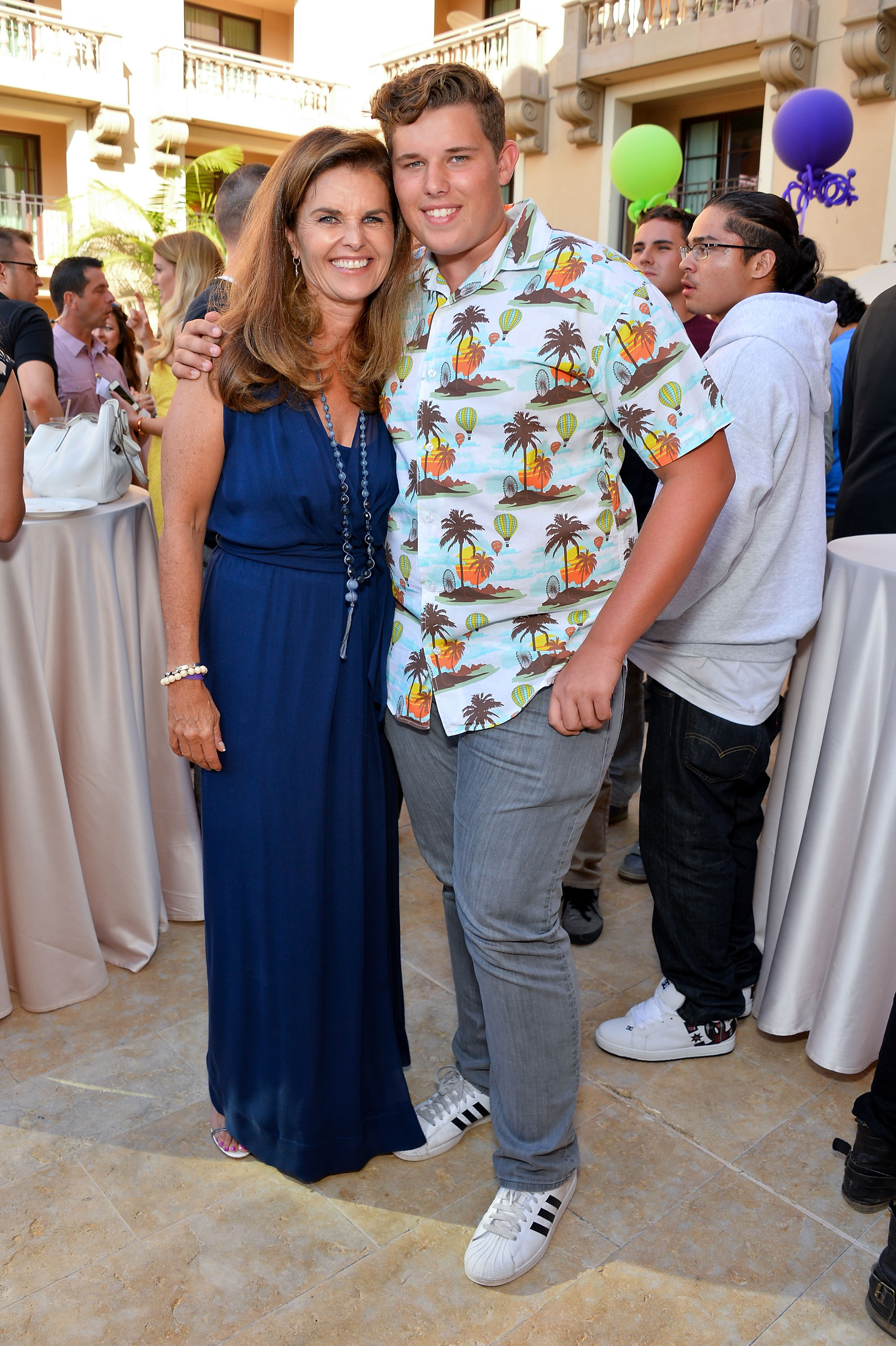 Maria Shriver und ihr Sohn Christopher teilen einen herzlichen Moment bei der gleichen Veranstaltung. Shriver trägt ein tiefblaues Kleid mit mehrlagigen Ketten, während Christopher sein typisches bedrucktes Hemd und eine graue Jeans trägt. Sie stehen eng beieinander, umgeben von Gästen in einer festlichen Atmosphäre.