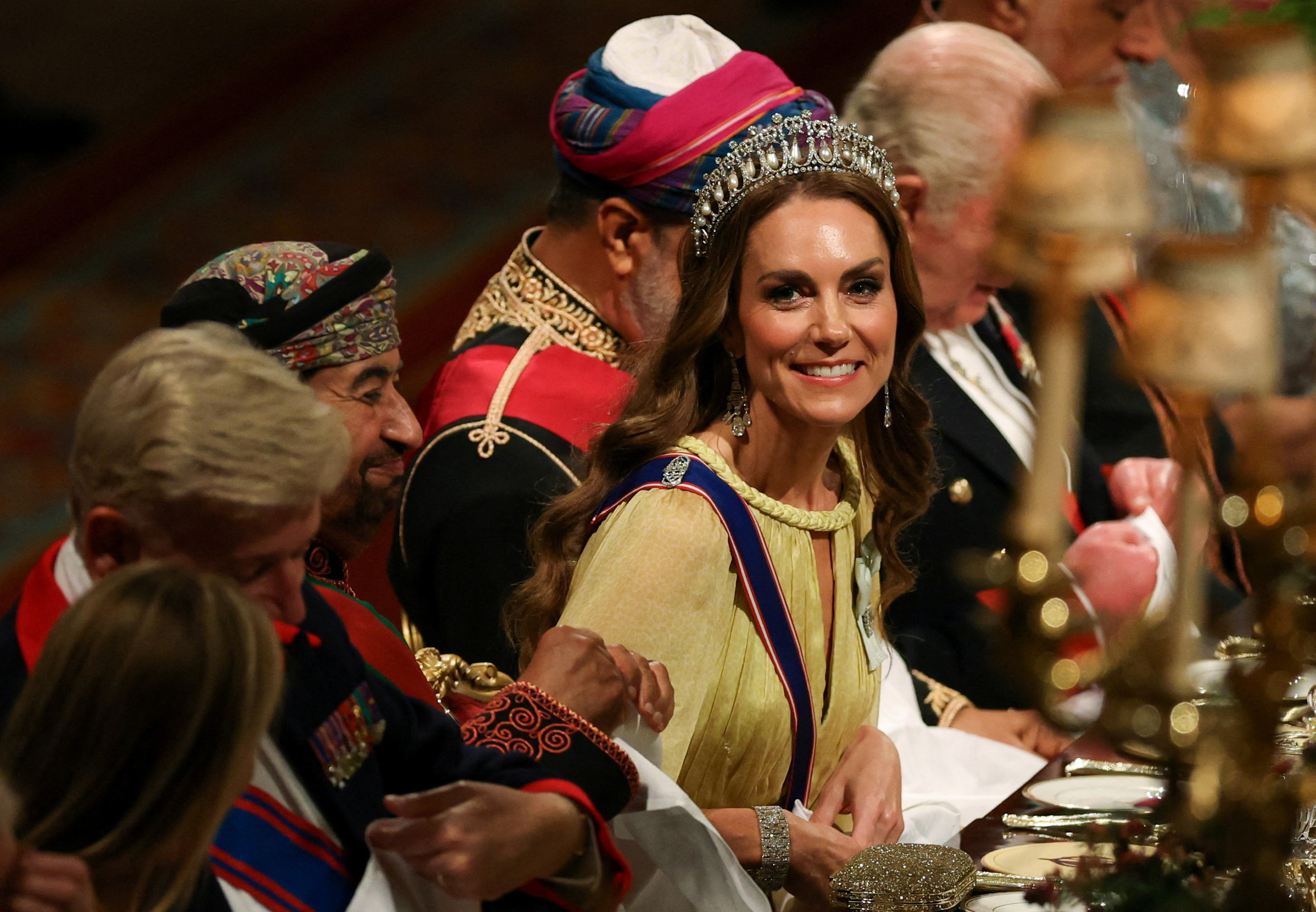 Catherine, Prinzessin von Wales, sitzt am 20. Oktober 2026 am Festtisch in der St. George’s Hall und strahlt herzlich, als das Abendessen beginnt. Ihr strahlender Beauty-Look besticht durch sanfte, brünette Wellen, die unter der mit Perlen und Diamanten besetzten „Lover’s Knot“-Tiara über ihre Schultern fallen, kombiniert mit diamantbesetzten Kronleuchter-Ohrringen, Smokey-Eyes und dem geflochtenen Seilausschnitt ihres gelben Cape-Kleides. | Quelle: Getty Images