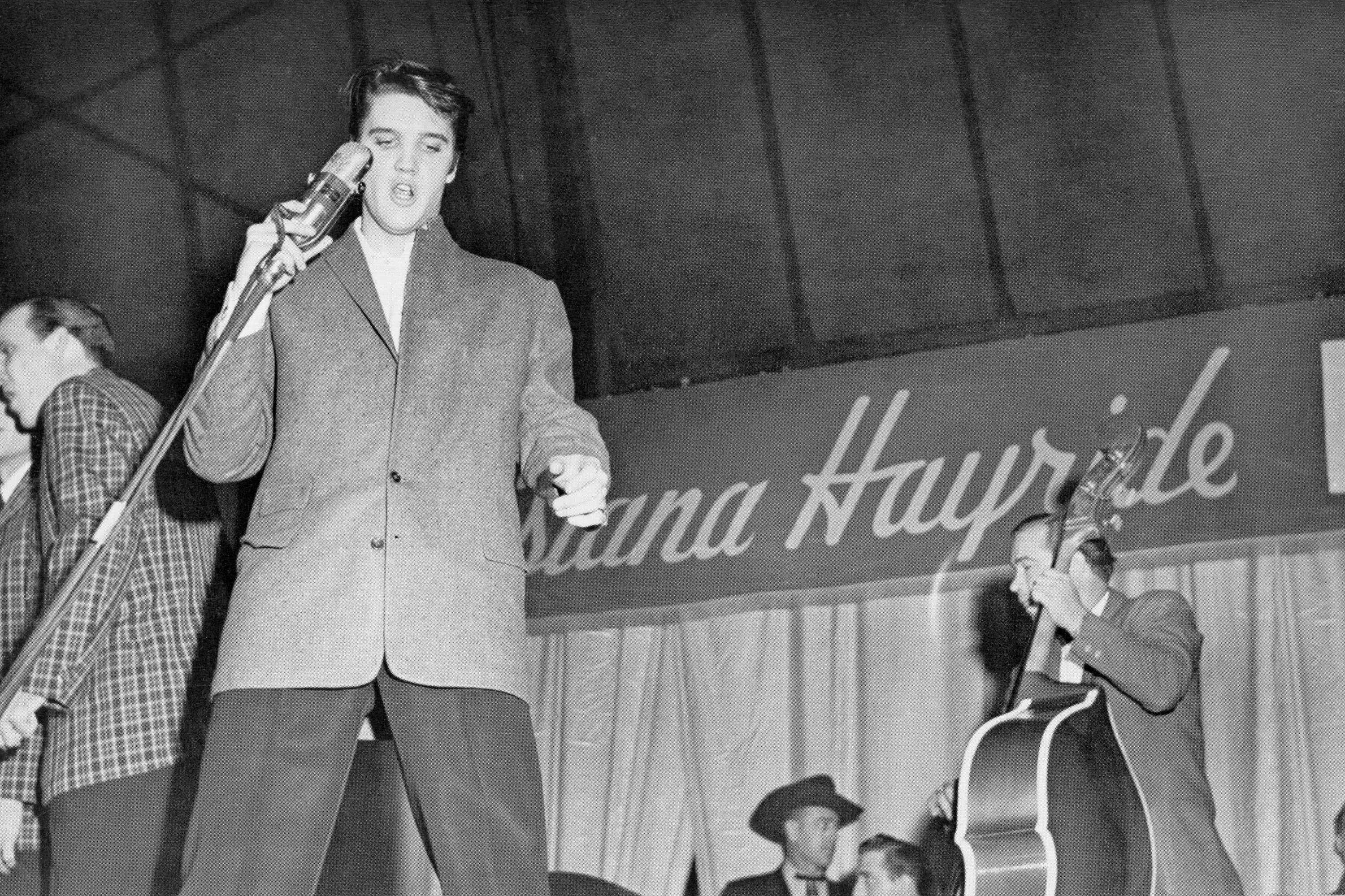 Der Rock-’n’-Roll-Sänger Elvis Presley singt 1954 während einer Tournee mit der Show „Louisiana Hayride“ zusammen mit dem Bassisten Bill Black in ein Vintage-Mikrofon. Er trägt ein maßgeschneidertes, einreihiges Tweed-Sakko über einer dunklen Hose und einem strahlend weißen Hemd mit Kragen. | Quelle: Getty Images