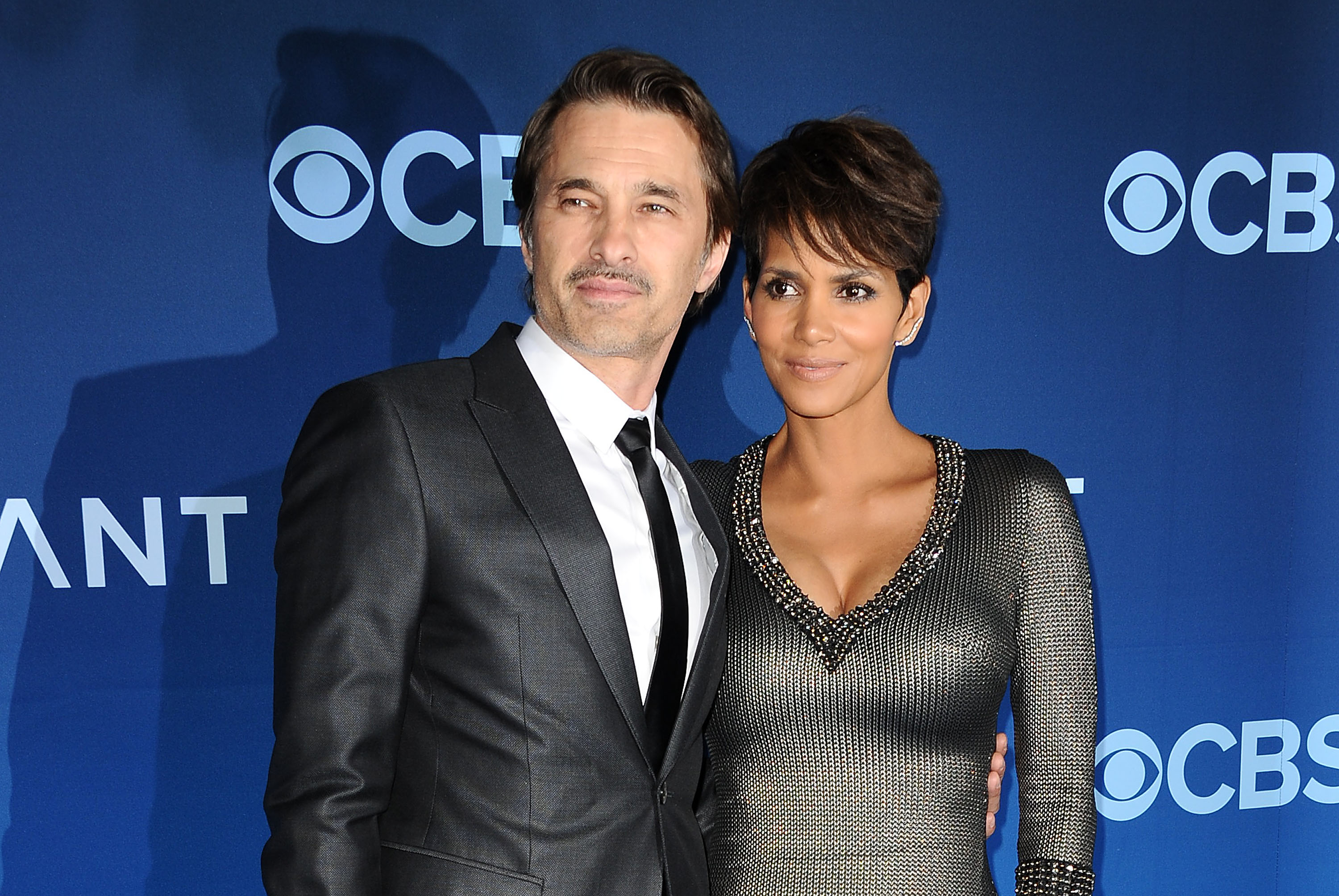 Der Schauspieler Olivier Martinez und die Schauspielerin Halle Berry posierten gemeinsam auf dem roten Teppich bei der Premiere von „Extant“, die am 16. Juni 2014 im California Science Center in Los Angeles, Kalifornien, stattfand. Martinez sah in einem dunkelgrauen, maßgeschneiderten Anzug, einem strahlend weißen Hemd und einer schmalen schwarzen Krawatte schick aus, während Berry ihn in einem metallisch strukturierten, gunmetalfarbenen Strickkleid mit funkelndem, perlenbesetztem Ausschnitt und ihrem typischen kurzen Pixie-Cut perfekt ergänzte. | Quelle: Getty Images