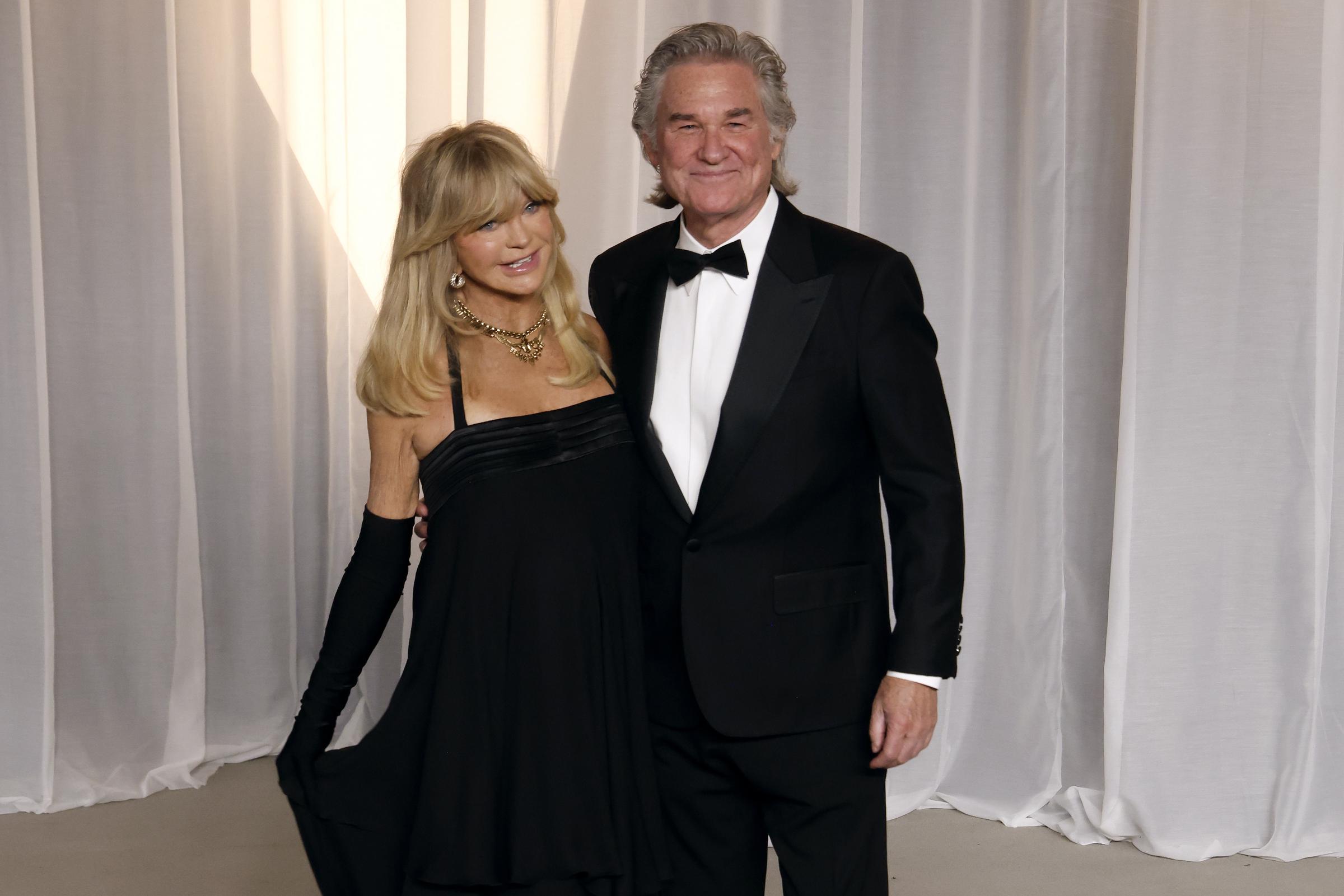 Goldie Hawn und Kurt Russell bilden in ihren aufeinander abgestimmten schwarzen Abendgarderoben ein mühelos schickes Paar. Hawn bezaubert in einem schwarzen Cocktailkleid mit Empire-Taille, kombiniert mit passenden opernlangen Handschuhen, auffälligem Goldschmuck und ihrem typischen voluminösen blonden Pony. Russell ergänzt ihren klassischen Stil in einem eleganten schwarzen Smoking mit spitzen Satinrevers, einem strahlend weißen Hemd und einer traditionellen schwarzen Fliege.