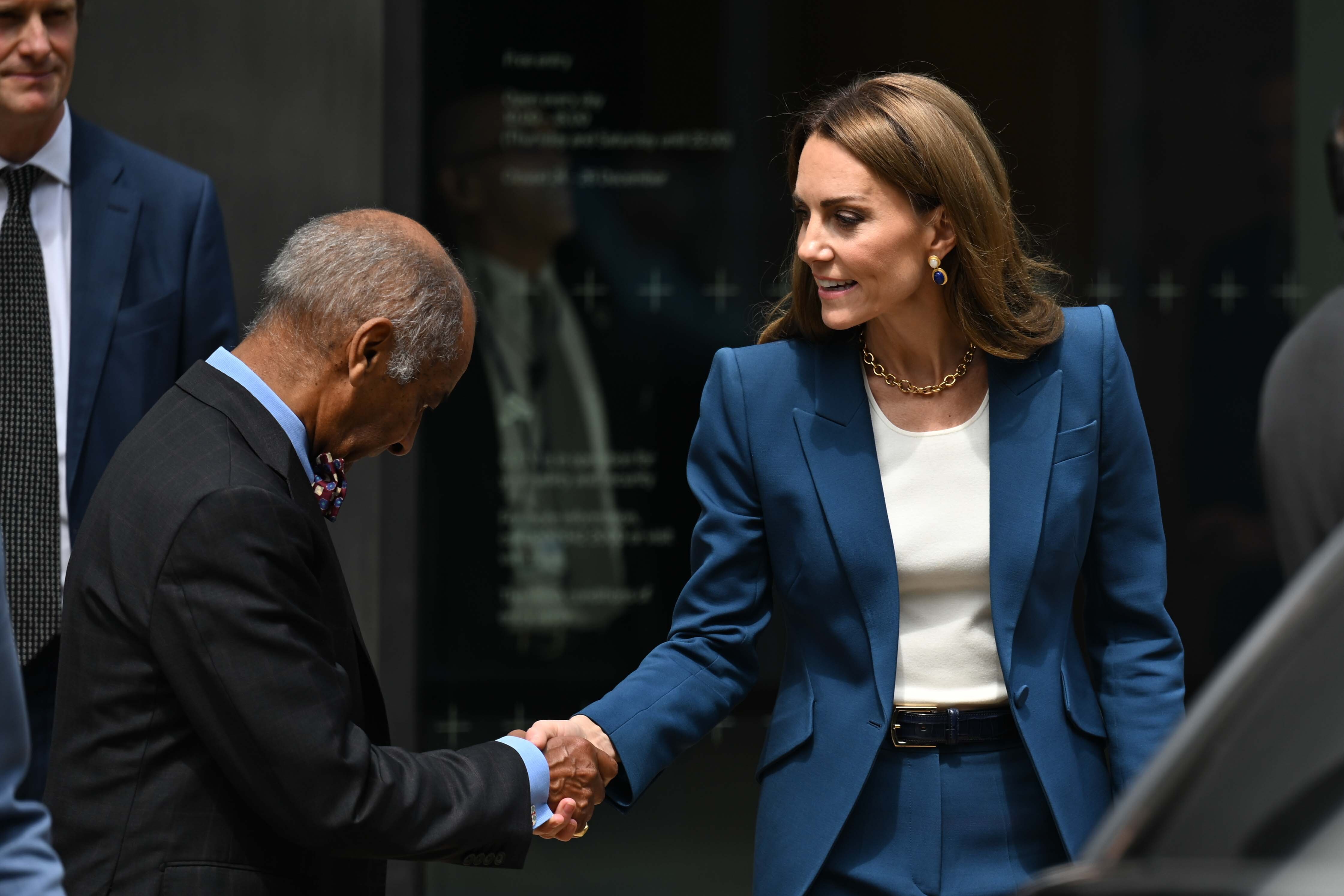 Mit einem höflichen Nicken und ständigem Augenkontakt begrüßt Prinzessin Catherine einen Beamten im V&A East Storehouse. Ihre Haltung ist anmutig, ihr Lächeln dezent, und ihre Saphir-Ohrringe setzen einen raffinierten Akzent zu ihrem Outfit.