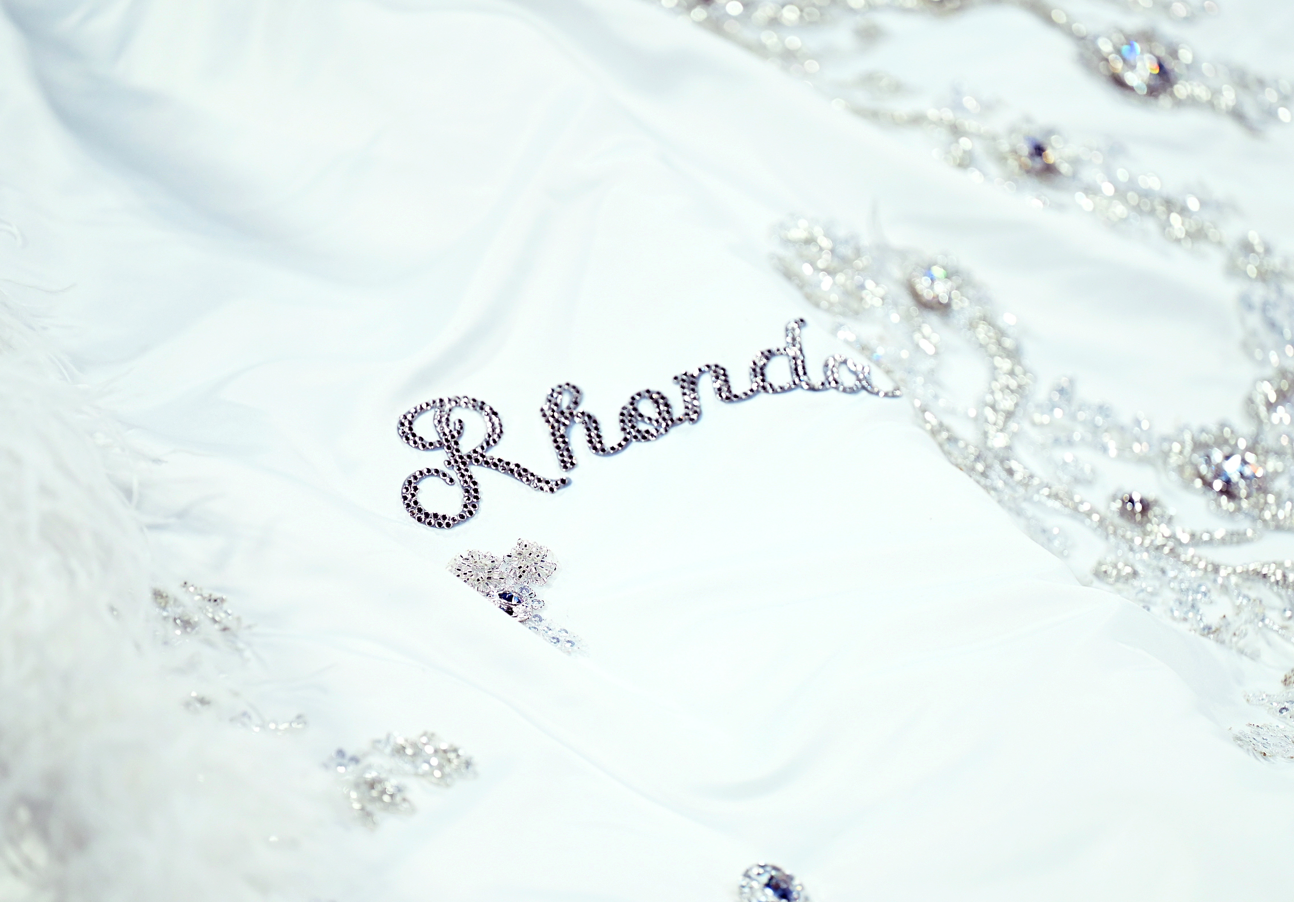 Eine Nahaufnahme der Schleppe lenkt die Aufmerksamkeit auf die Stickerei, bei der der Name „Rhonda“ inmitten der Details deutlich sichtbar ist. Die übrigen Namen, die ihrer Kinder und Enkelkinder, schimmern durch und bieten flüchtige Einblicke, die das herzliche Design des Kleides unterstreichen.
