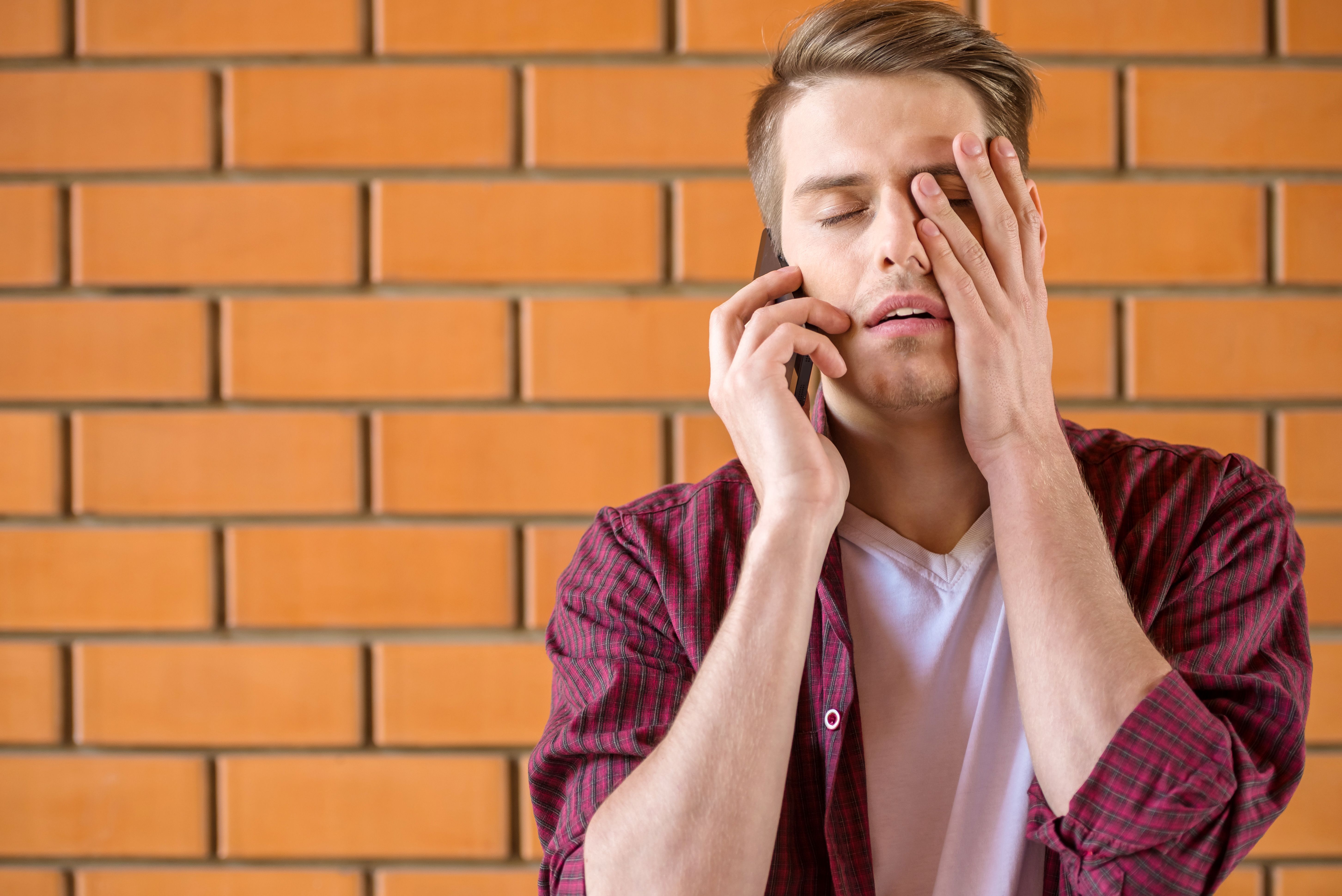 Ein junger Mann, verzweifelt am Telefon, hinter ihm eine Backsteinwand. | Quelle: Shutterstock