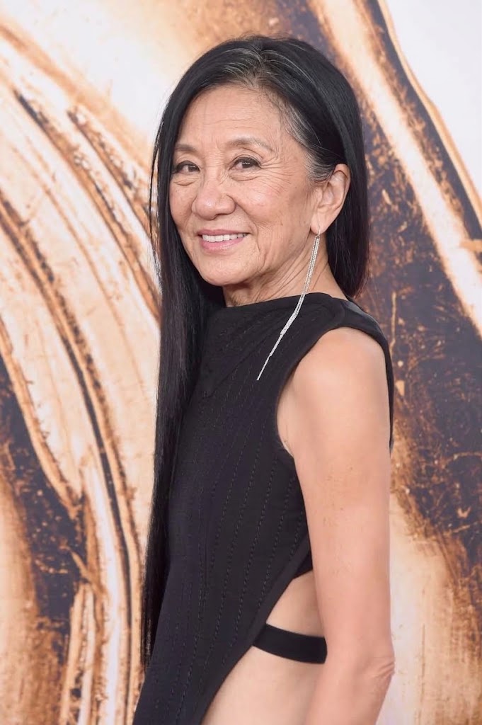 Diese KI-Darstellung zeigt Vera Wang ganz im Zeichen eines natürlichen Alterungsprozesses – mit zarten Krähenfüßen, Lachfalten und feinen silbernen Strähnen, die sich in ihr glattes, seitlich geflochtenes Haar schlängeln. Vor einem wirbelnden, neutralen Hintergrund, der an den roten Teppich bei High-Fashion-Preisverleihungen erinnert, strahlt die simulierte Designerin in einem ärmellosen schwarzen Kleid mit dramatisch seitlich offenem Schnitt und einem langen, funkelnden Kristall-Ohrring. | Quelle: Gemini