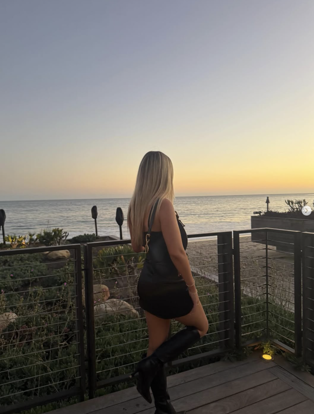 Finley Lockwood genießt den Sonnenuntergang auf einer Terrasse direkt am Meer. Auf dem Foto ist sie von hinten zu sehen – in einem eleganten schwarzen, ärmellosen Minikleid und kniehohen schwarzen Lederstiefeln, während ihr langes blondes Haar ihr über den Rücken fällt. | Quelle: Instagram/finleyl.ockwood