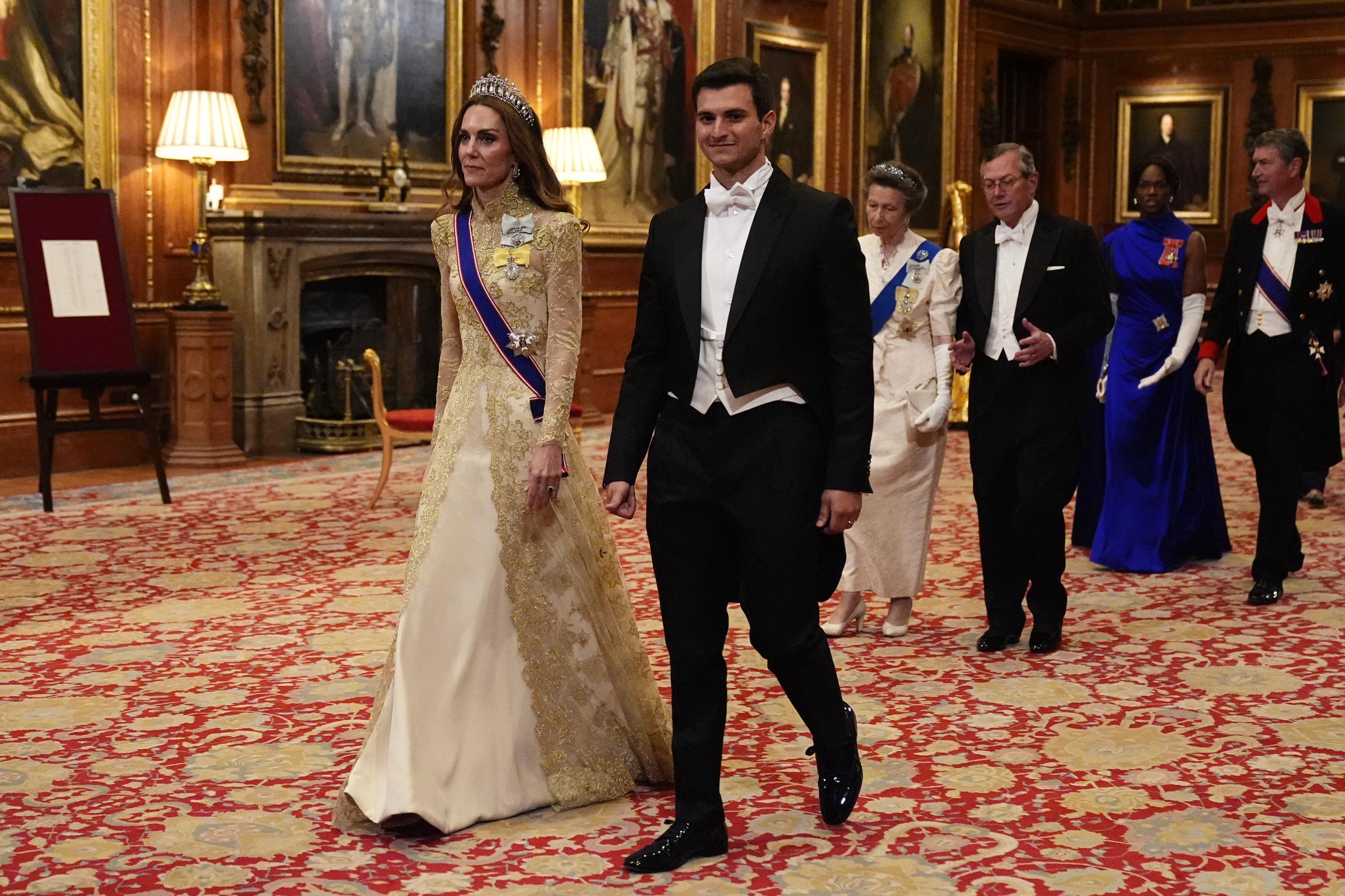 Catherine, Prinzessin von Wales, schreitet am 17. September 2025 an der Seite von Michael Boulos während des feierlichen Einzugs zum Staatsbankett im Windsor Castle, das zu Ehren des US-Präsidenten Donald Trump und der First Lady Melania Trump stattfindet. Catherine sieht umwerfend aus in einem elfenbeinfarbenen und goldenen Spitzenkleid mit langen Illusionsärmeln und aufwendigen Goldstickereien, ergänzt durch Königin Marys „Lover’s Knot“-Tiara, die Schärpe des Royal Victorian Order und den Royal Family Order von König Charles III., während Boulos in einem schwarzen Frack und einer weißen Krawatte elegant gekleidet ist. | Quelle: Getty Images