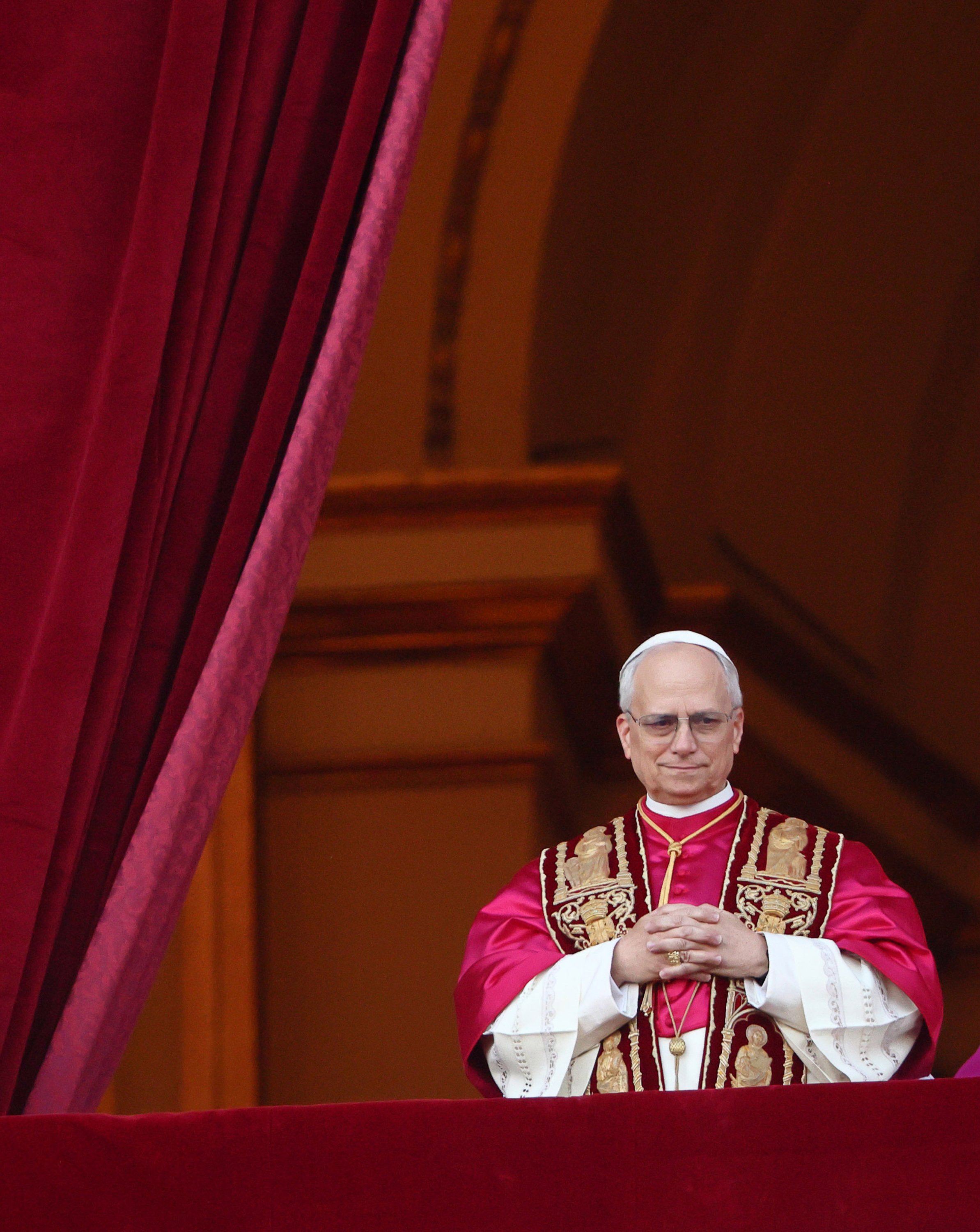 Papst Leo XIV. steht feierlich zwischen reichen Samtvorhängen, die Hände über den mit goldenen Heiligen bestickten purpurroten Gewändern gefaltet