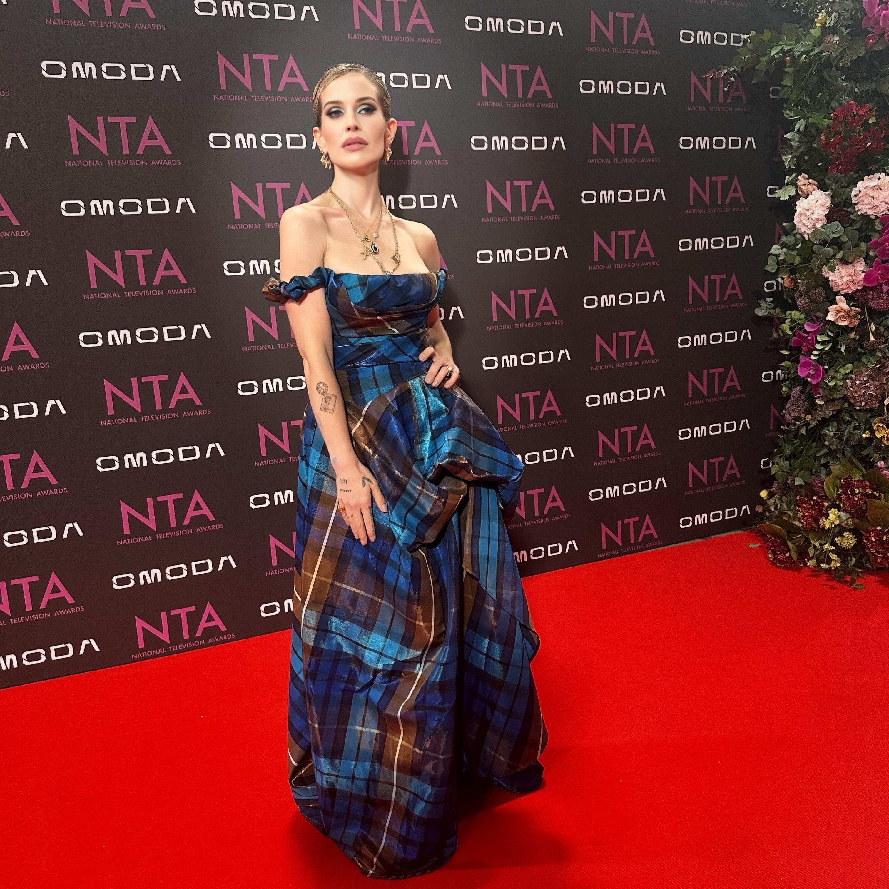 Kelly präsentiert die asymmetrische Silhouette und den weiten Rock ihres 3.485 € teuren Tartan-Kleids von Vivienne Westwood vor dem offiziellen NTA-Hintergrund. | Quelle: Instagram/kellyosbourne
