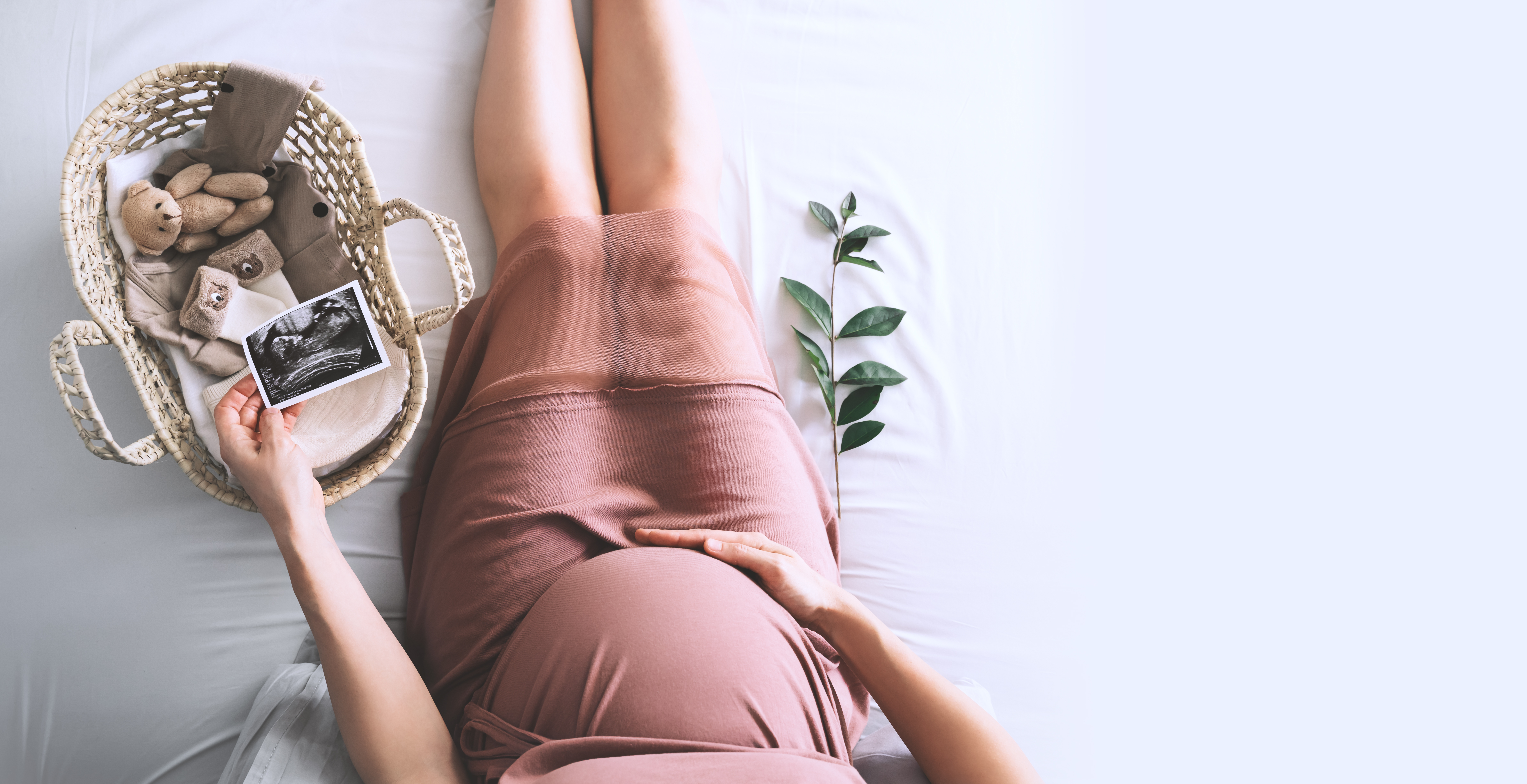 Eine Frau hält einen Scan ihres Babys in der einen und ihren schwangeren Bauch in der anderen Hand neben einem Korb mit Babyzubehör | Quelle: Shutterstock