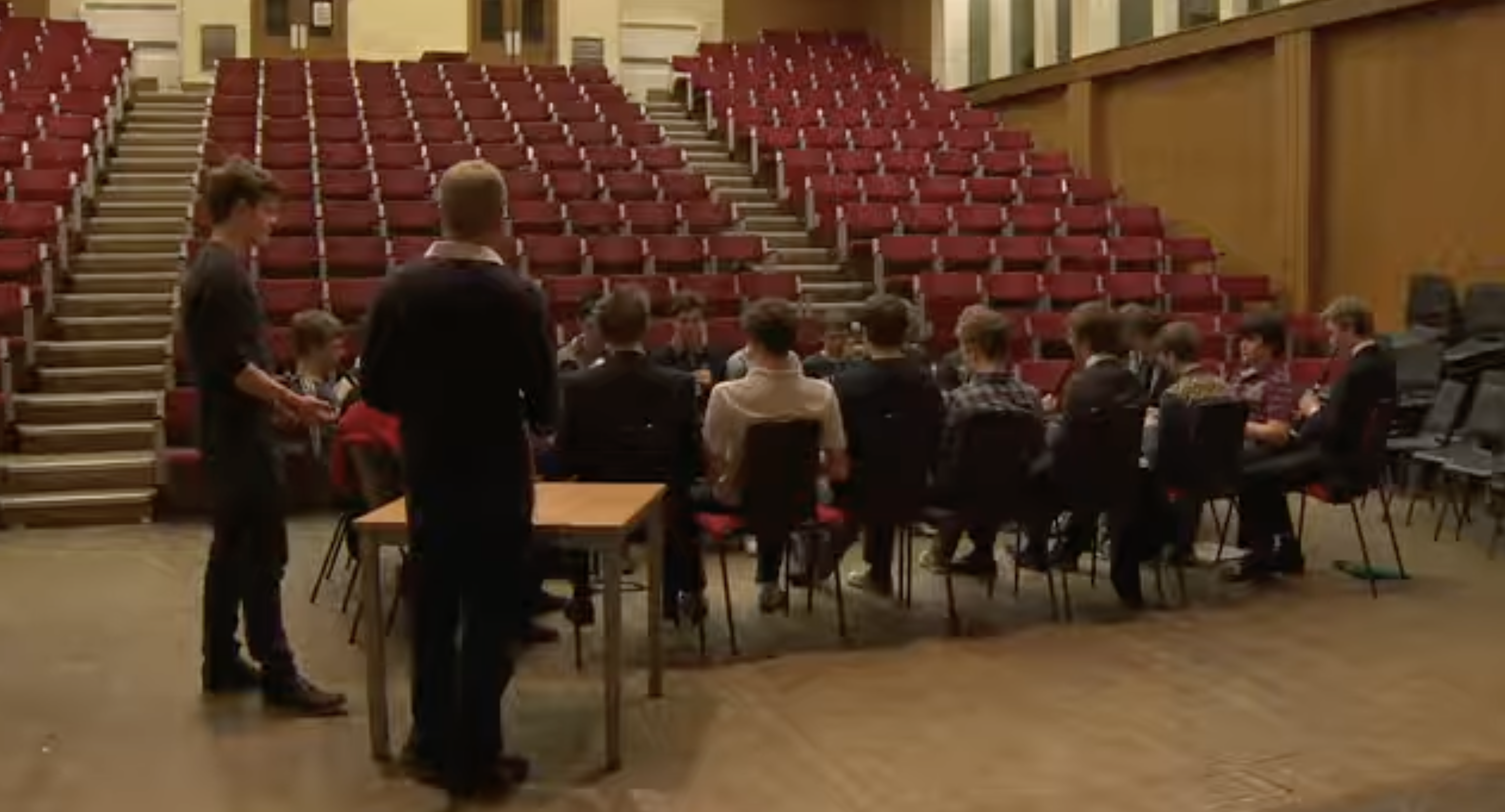 Die Schüler sitzen in einer Gruppe auf dem Boden einer großen Halle, die von Reihen roter, stufenförmiger Sitzplätze umgeben ist. Die Versammlung gibt einen Einblick in die Gemeinschaftsräume, die am Eton College für Aktivitäten genutzt werden. | Quelle: YouTube/Paradoxyy