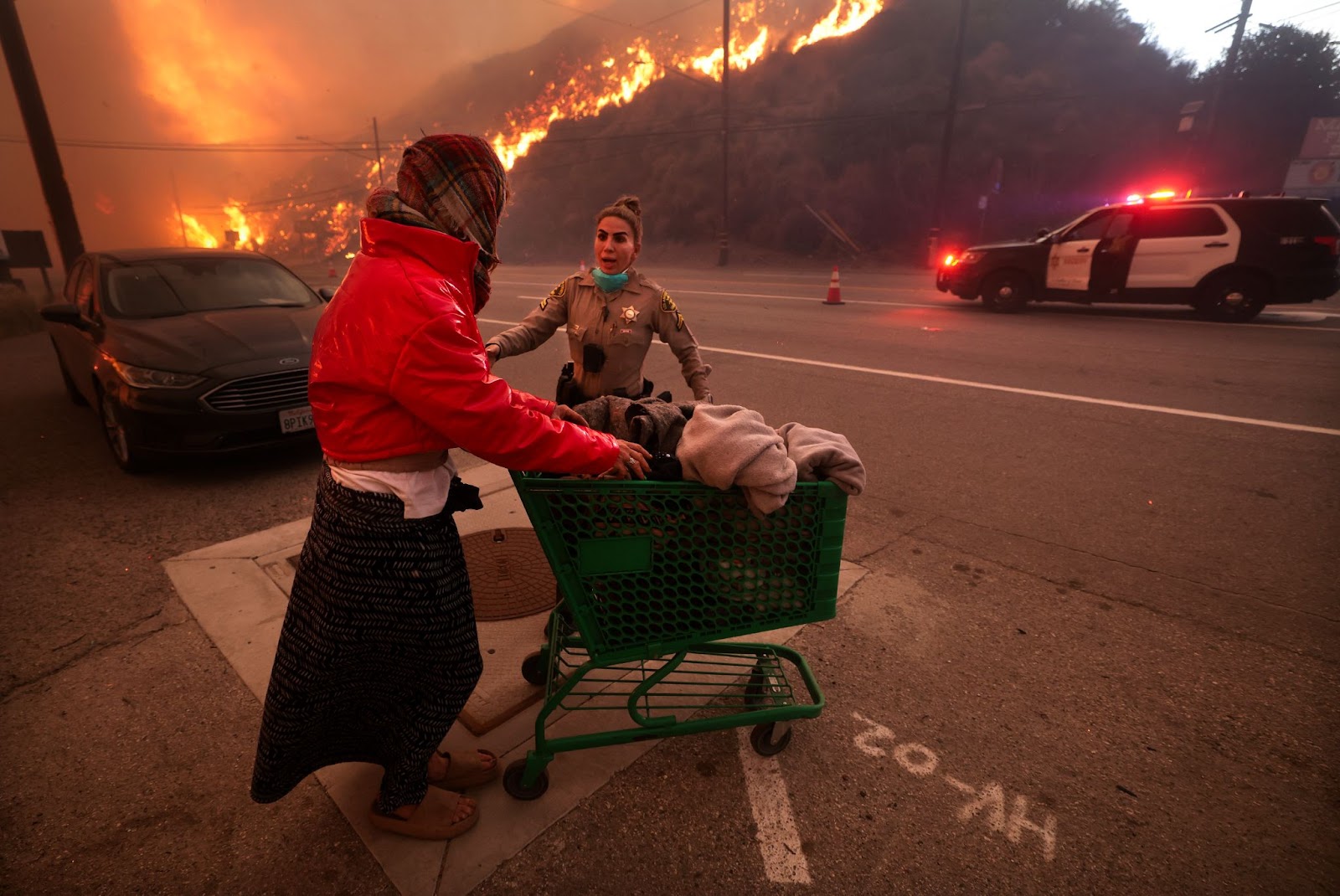 Eine Polizistin eskortiert eine obdachlose Frau vom Pacific Coast Highway und dem Topanga Canyon Boulevard weg, während das Palisades-Feuer in Los Angeles, Kalifornien, am Dienstag, 7. Januar 2025, wütet. | Quelle: Getty Images