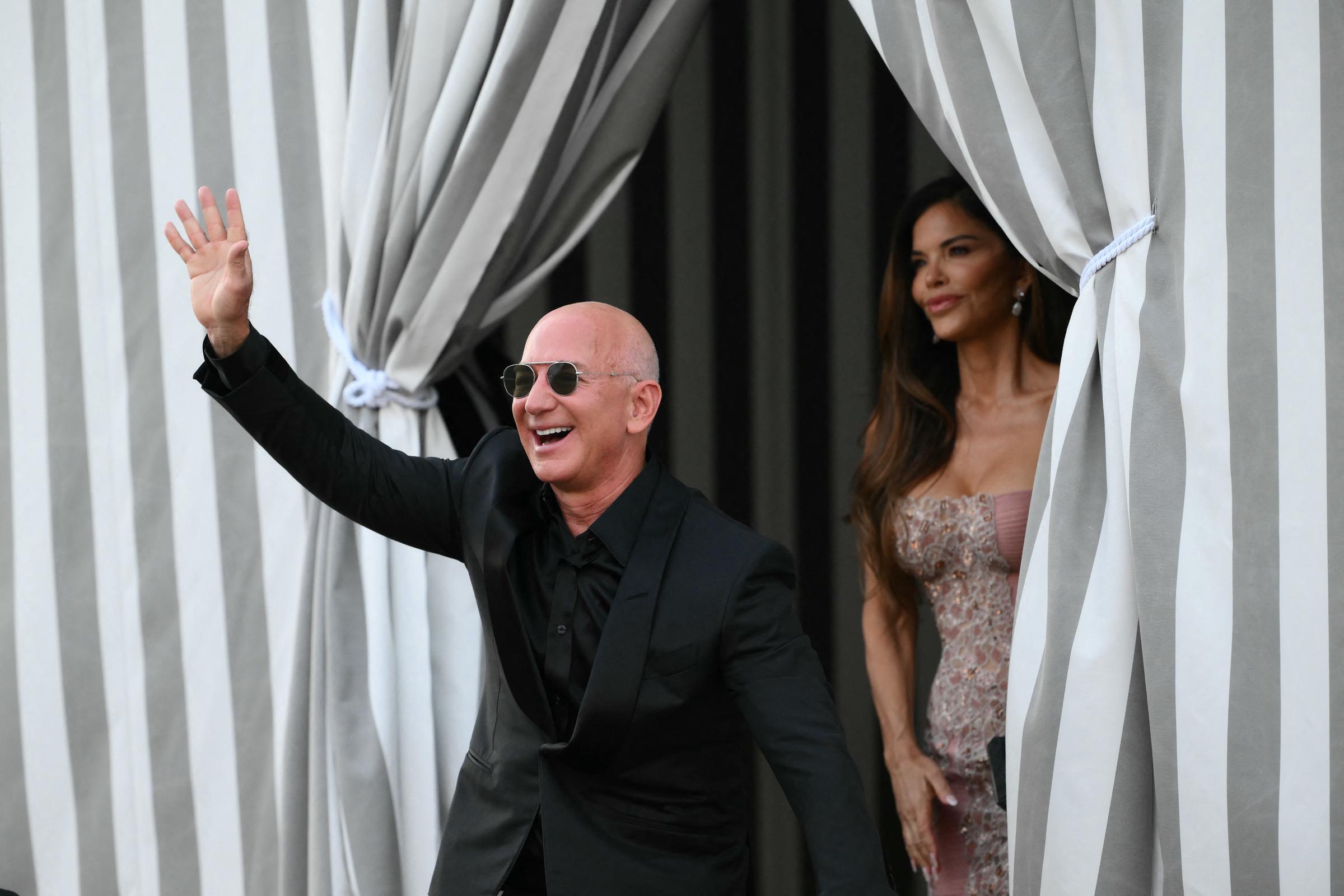 Jeff Bezos strahlt und winkt den Schaulustigen energisch zu. Er trägt einen scharfen schwarzen Anzug und eine Piloten-Sonnenbrille, während Lauren Sánchez anmutig hinter ihm steht, in einem schimmernden, erröteten Kleid mit aufwändigen Perlen, ihr langes Haar in lockeren Wellen gestylt, während sie zwischen eleganten gestreiften Vorhängen hervorschaut.