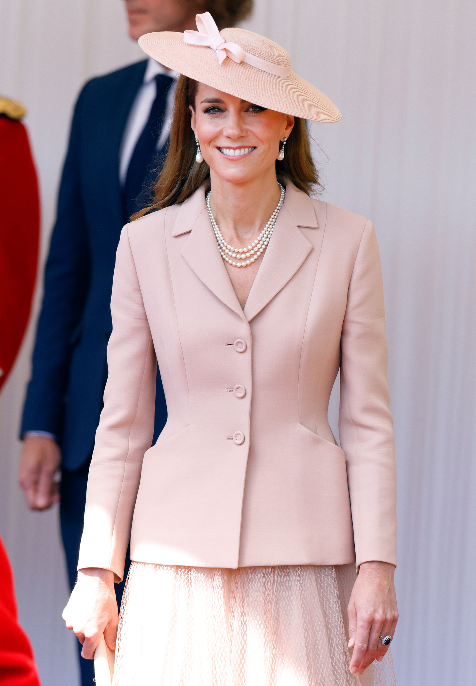 Catherine, Prinzessin von Wales, lächelt herzlich vom königlichen Podium an der Datchet Road in Windsor, England, während der feierlichen Begrüßungszeremonie für den französischen Präsidenten Emmanuel Macron und Brigitte Macron am 8. Juli 2025. Als Hommage an die französische Couture-Diplomatie trägt Catherine eine blassrosa, maßgeschneiderte Barjacke von Christian Dior über einem passenden Plissee-Tüllrock, wunderschön ergänzt durch Königin Elizabeth II.s dreisträngige Perlenkette, Perlen-Ohrhänger und einen schräg sitzenden Untertassenhut, der mit einer zarten Schleife verziert ist. | Quelle: Getty Images
