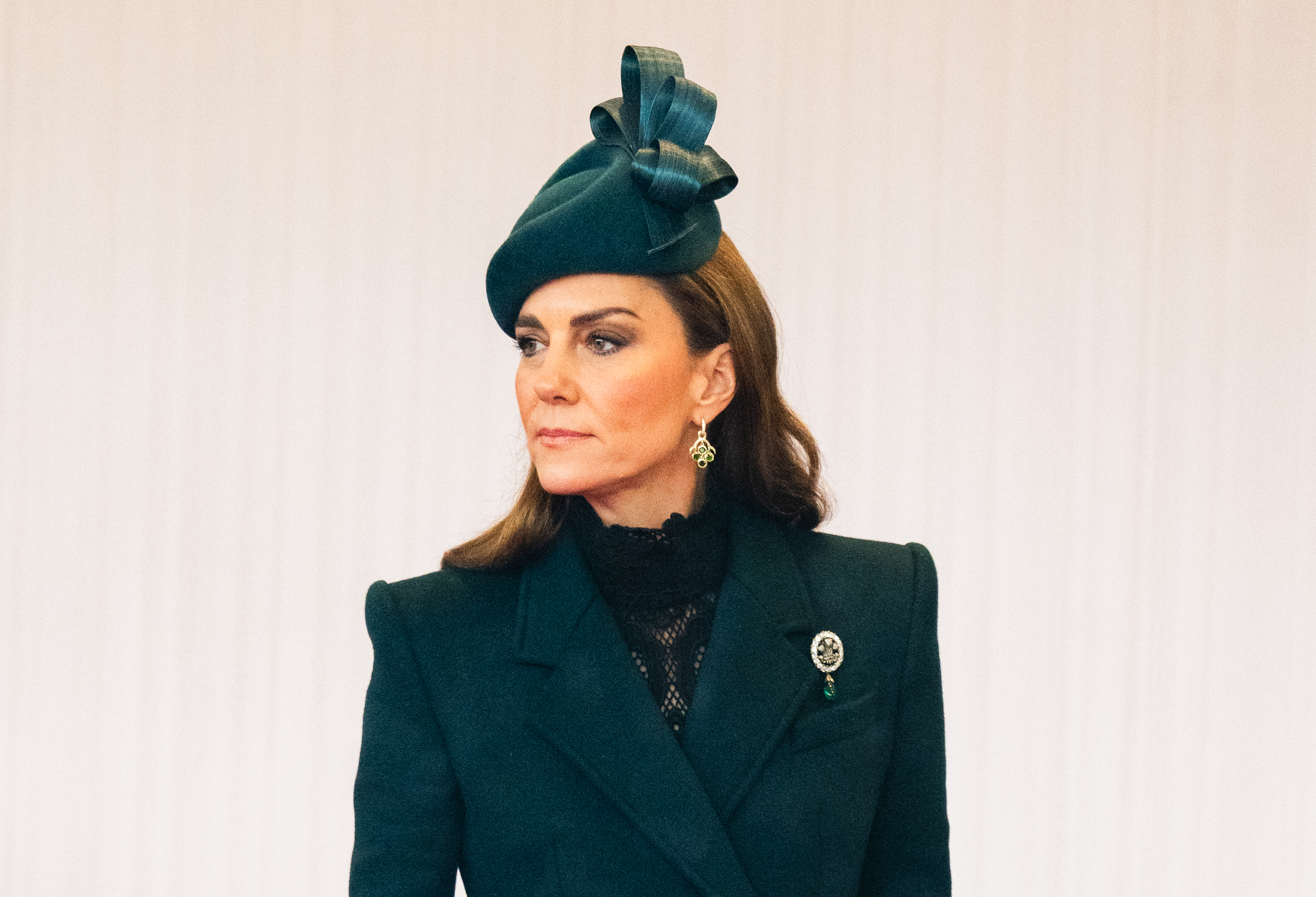 Ein Nahaufnahme-Porträt zeigt Catherine, Prinzessin von Wales, am 20. Oktober 2026 und verdeutlicht das aufwendige Styling ihres Tagesoutfits. Ihr maßgeschneiderter Mantel mit fallendem Revers umrahmt einen hohen Kragen aus transparenter schwarzer Spitze, dazu trägt sie einen skulpturalen grünen Fascinator mit Schleife, „Cassandra Goad“-Kronleuchter-Ohrringe und die funkelnde „Prince of Wales Feathers“-Brosche, die an ihrer linken Schulter befestigt ist. | Quelle: Getty Images