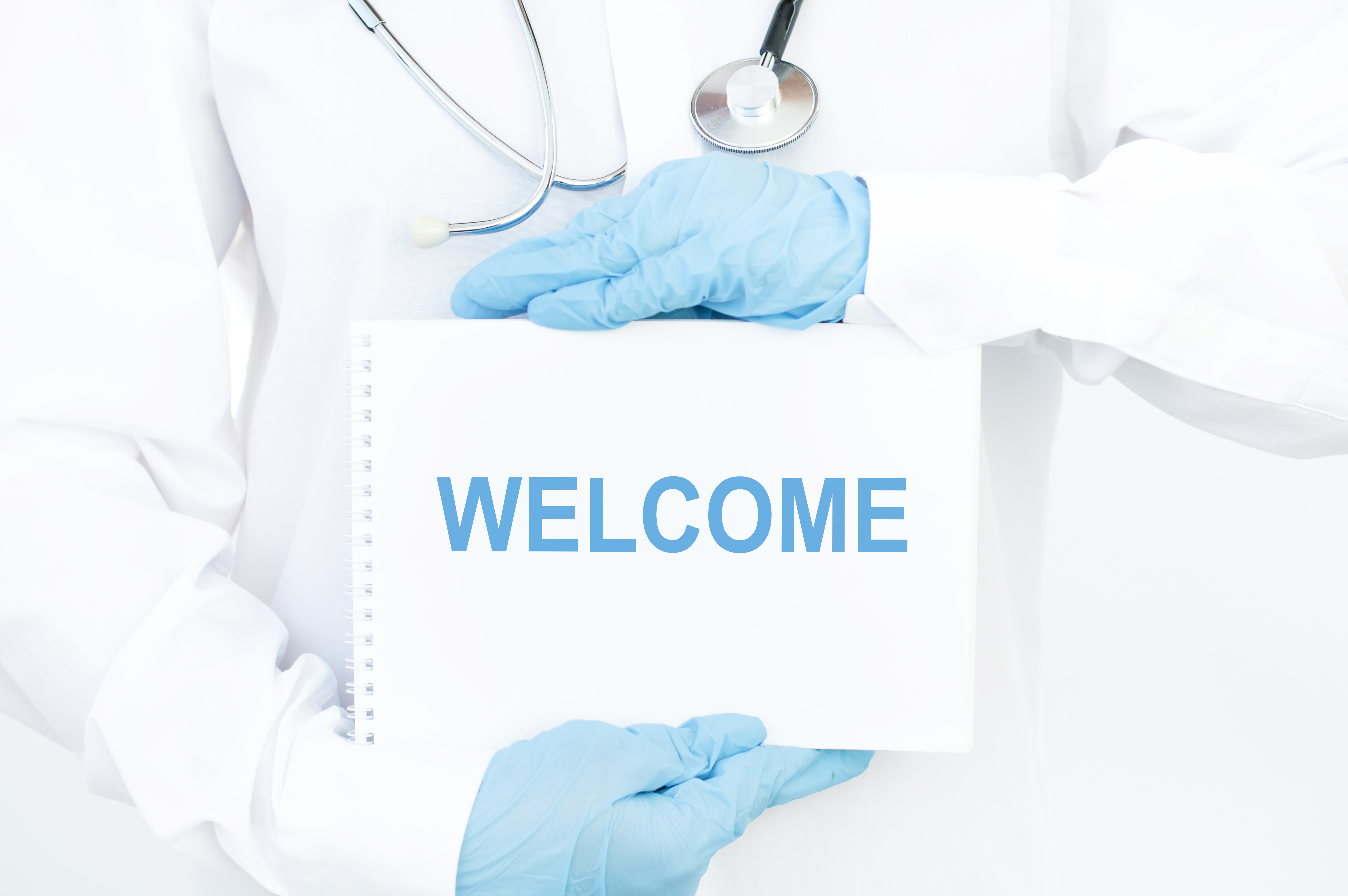 Arzt mit weißem Kittel und blauen Handschuhen hält ein "Welcome"-Schild. | Quelle: Shutterstock