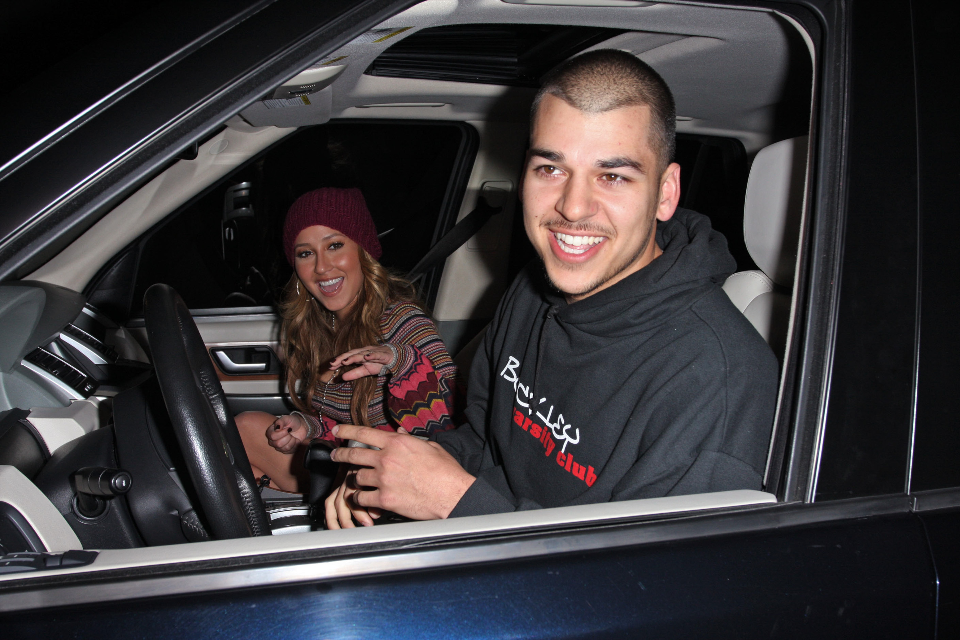 Rob Kardashian lächelt am Steuer, während eine Frau neben ihm den Fotografen bei einem nächtlichen Ausflug zuwinkt. Das Foto fängt einen entspannten, fröhlichen Moment aus seinen frühen Jahren im Rampenlicht ein. | Quelle: Getty Images.