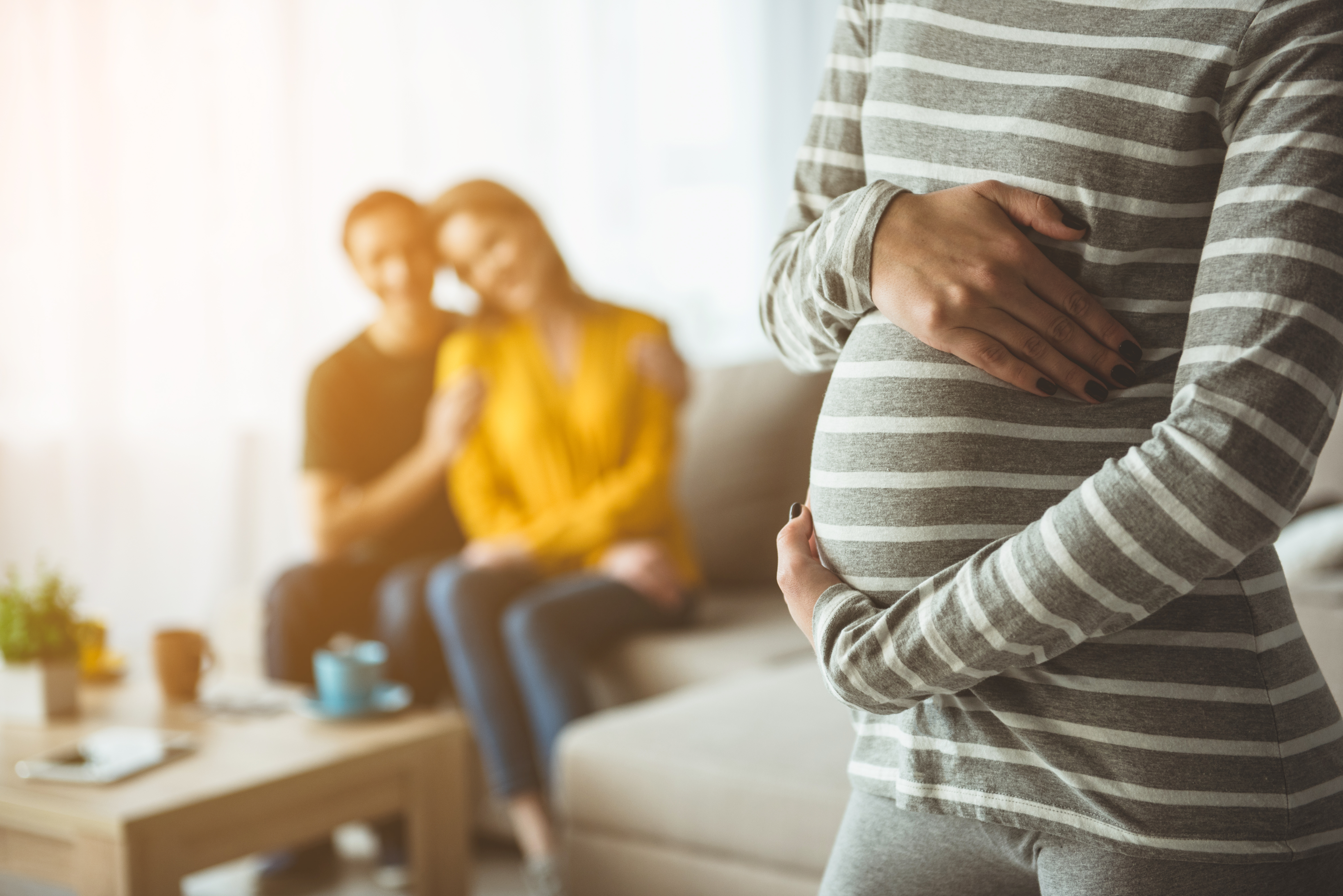 Eine schwangere Frau umfängt ihren Babybauch, im Hintergrund ist ein glückliches Paar zu sehen. | Quelle: Shutterstock