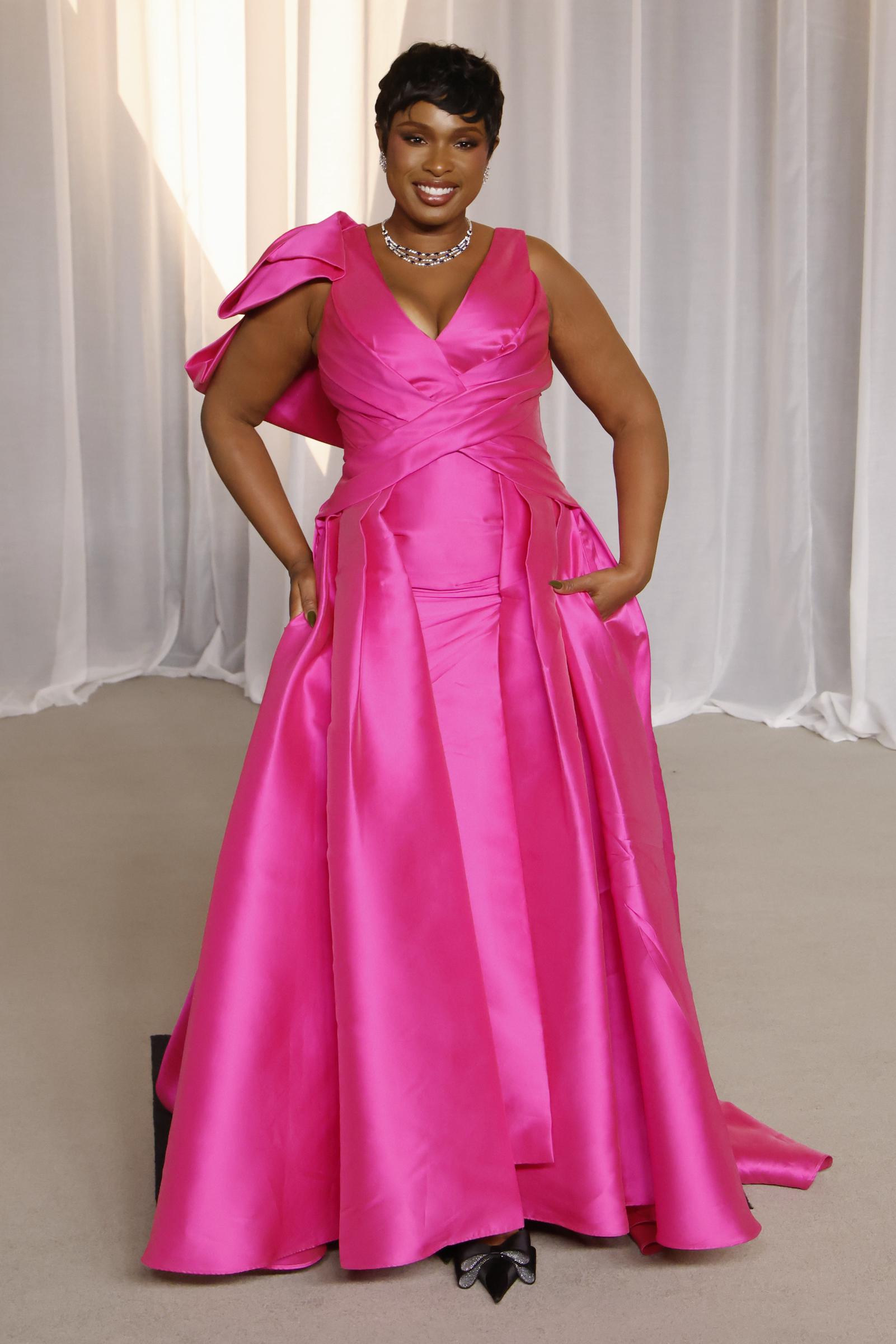 Jennifer Hudson zieht alle Blicke auf sich in einem leuchtend pinkfarbenen Satinkleid mit tiefem V-Ausschnitt und kunstvoll drapiertem Oberteil. Eine asymmetrische, voluminöse Schleife ziert eine Schulter, während der weite, bodenlange Rock mit eingenähten Taschen für mühelosen Charme sorgt. Die EGOT-Gewinnerin rundet den atemberaubenden Look mit einer funkelnden Diamant-Halskette, spitz zulaufenden Pumps mit Kristallschleifen und einem eleganten Pixie-Cut ab.