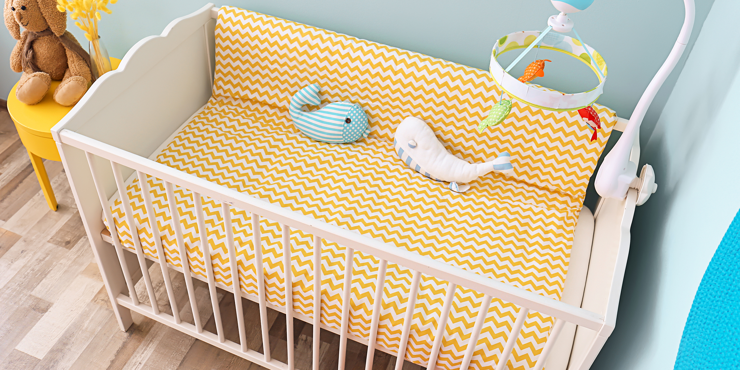 Ein Babybett mit gelber Chevron-Bettwäsche und Plüschtieren in einer hellen Kinderzimmerecke | Quelle: Shutterstock