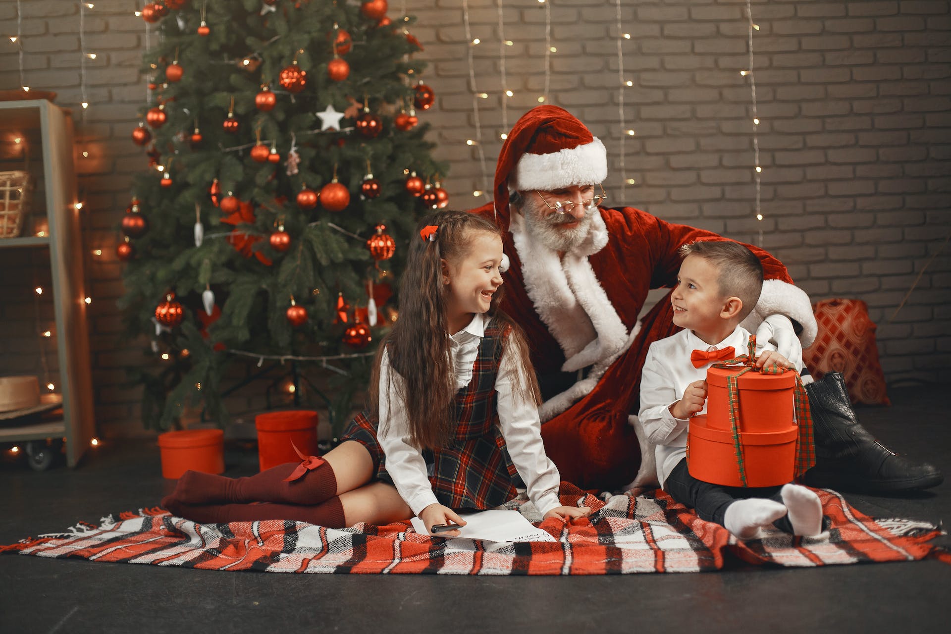 Mann im Weihnachtsmannkostüm sitzt mit einem Jungen und einem Mädchen neben einem Weihnachtsbaum | Quelle: Pexels